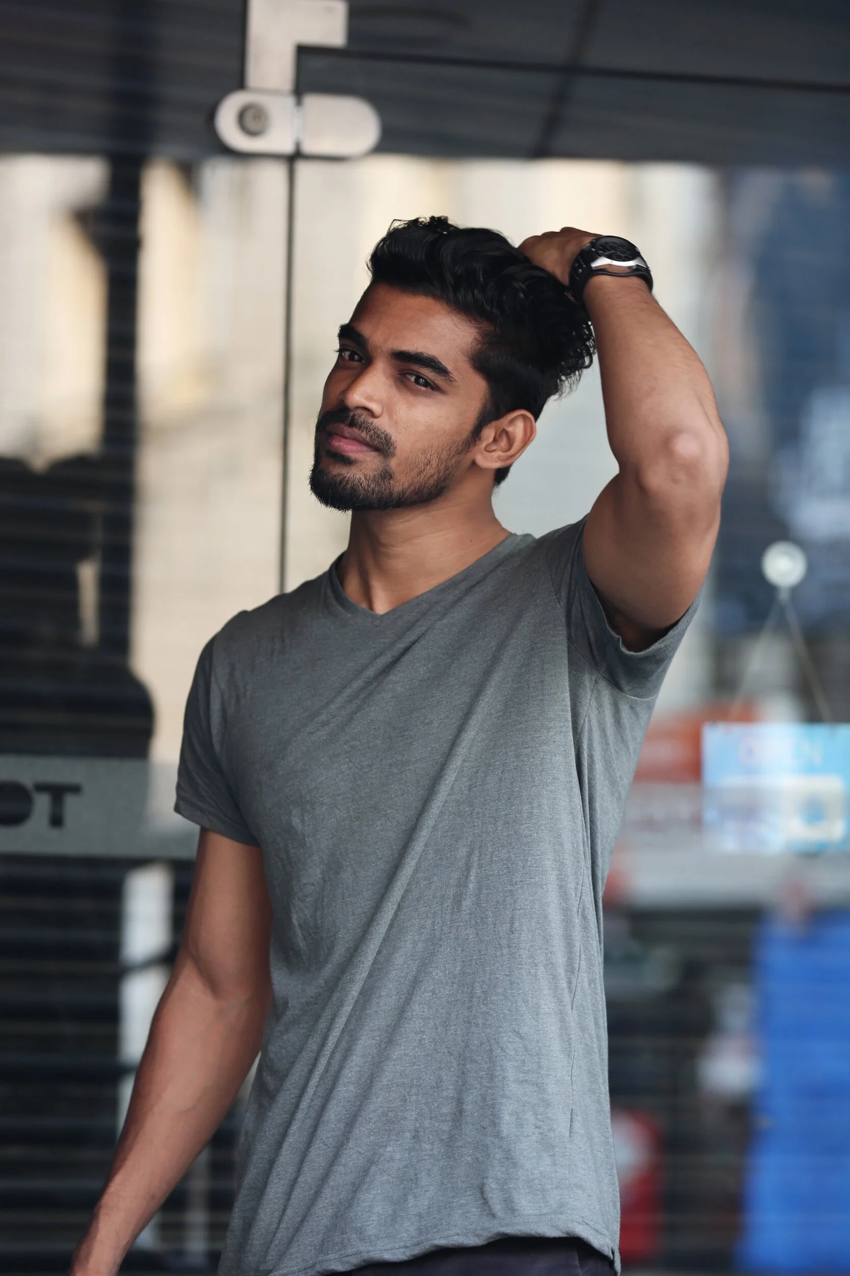 A young man with dark hair, a beard, wearing a gray t-shirt and a black watch, standing against a glass wall, touching his hair and looking into the camera.
