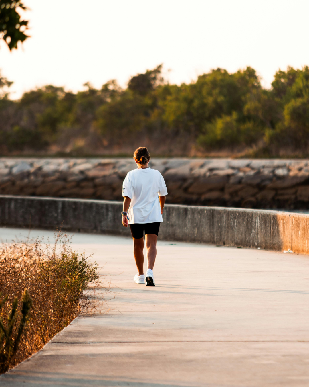 A woman walking on a paved path near a body of water, with trees in the background during sunset.