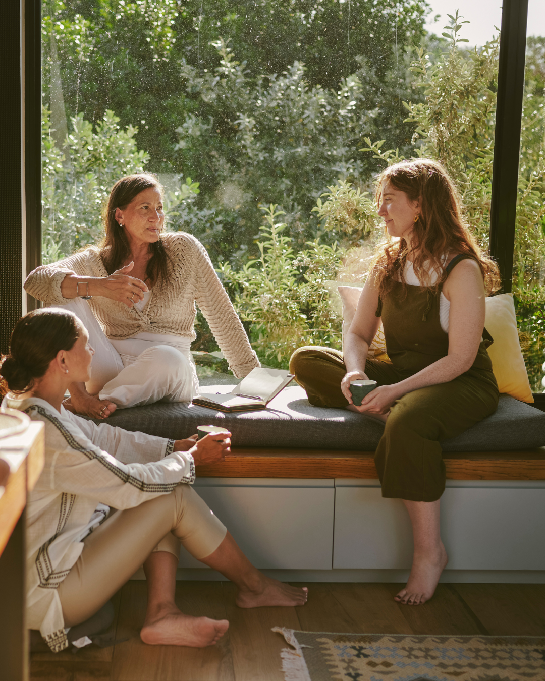 Four women having a conversation in a sunlit room with large windows and greenery outside. Three women are sitting on the floor and one on a window seat, all with cups or a book.
