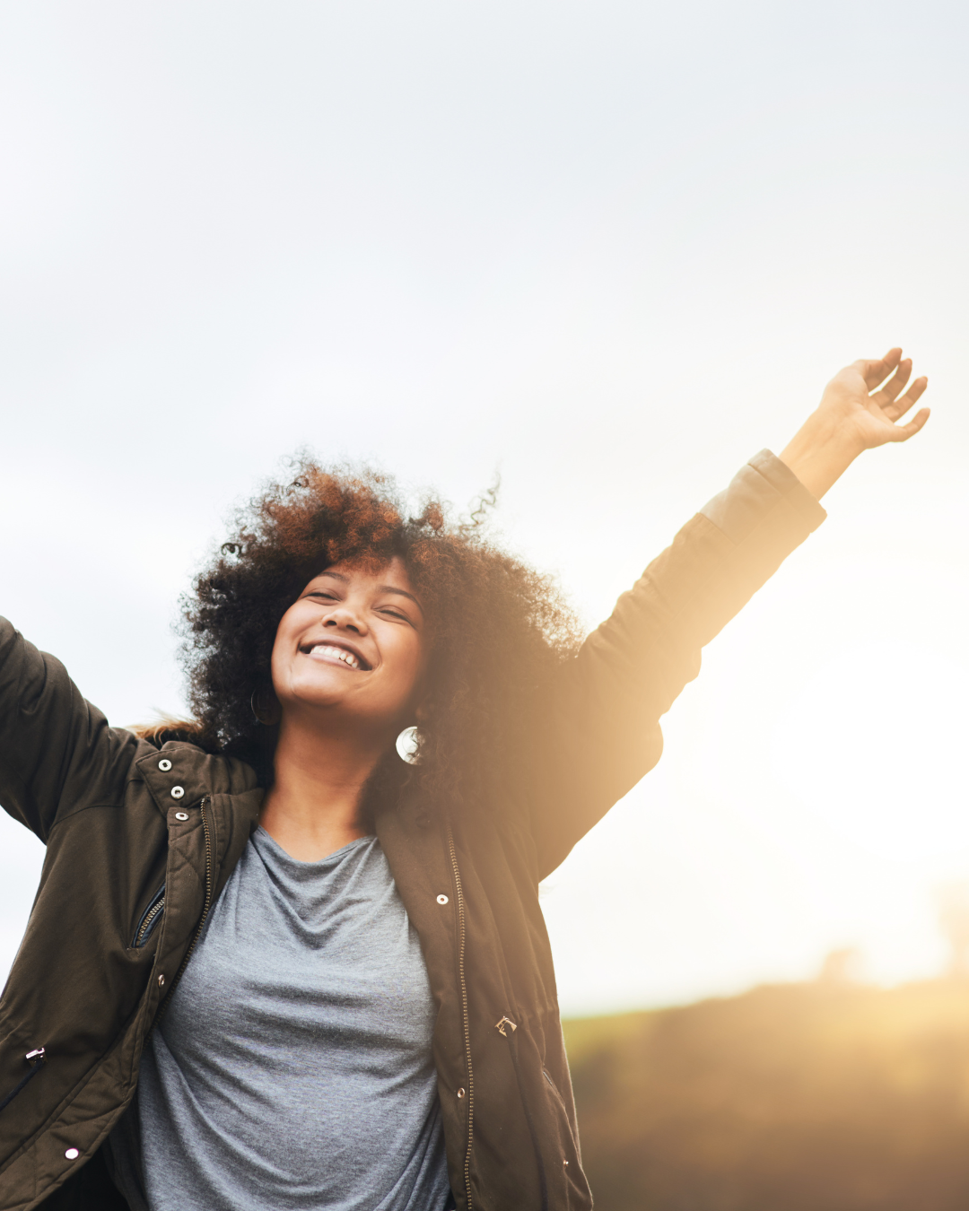 A joyful woman with curly hair standing outdoors with arms raised, enjoying sunlight.