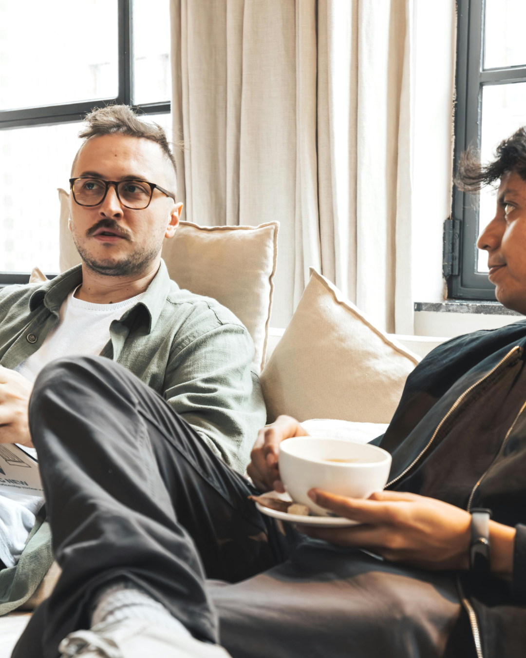 Two people sitting on a sofa in a bright room, having a conversation. One person is holding a cup of coffee or tea.