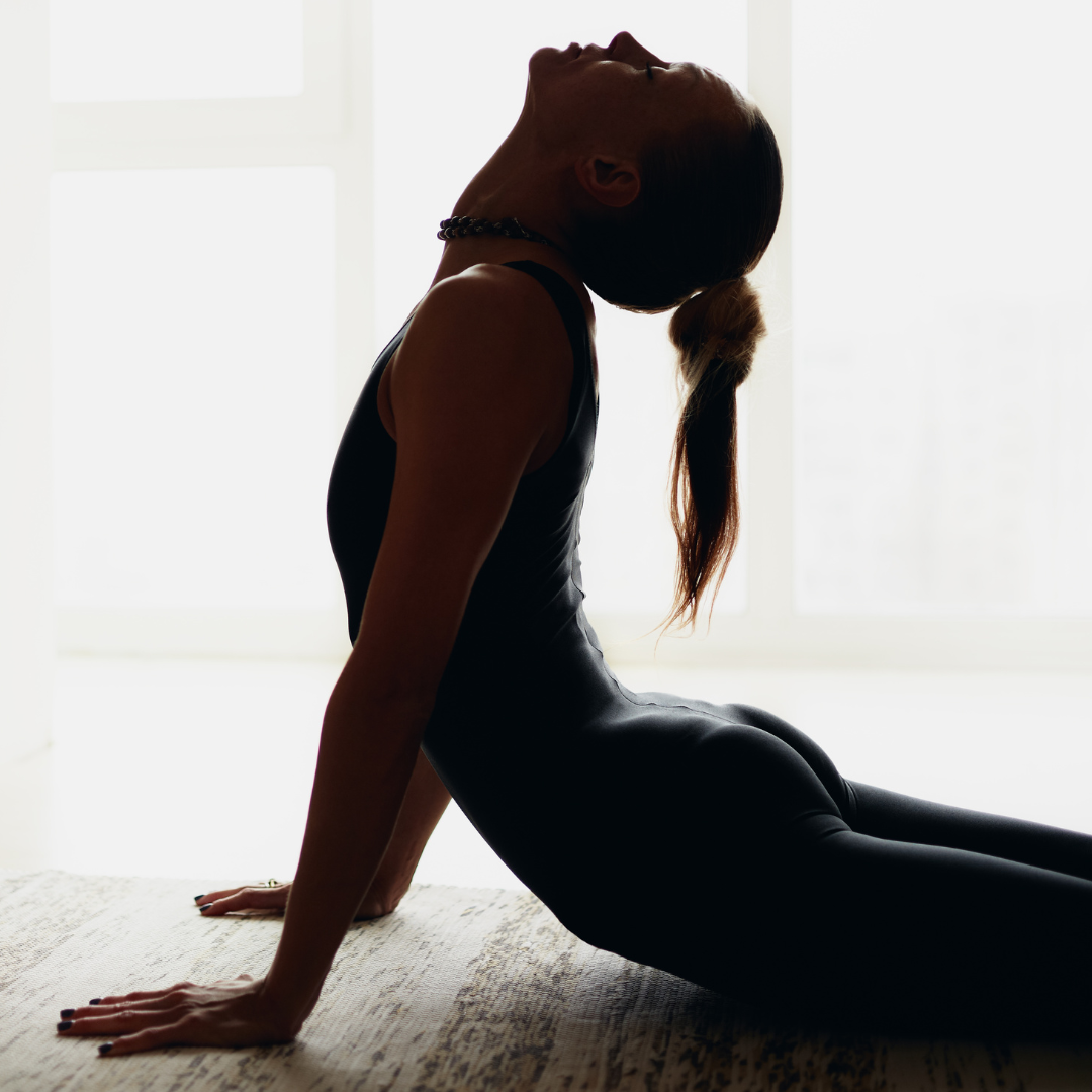 A woman practicing yoga indoors in front of large windows, sitting back with her hands on the floor and her head tilted back.