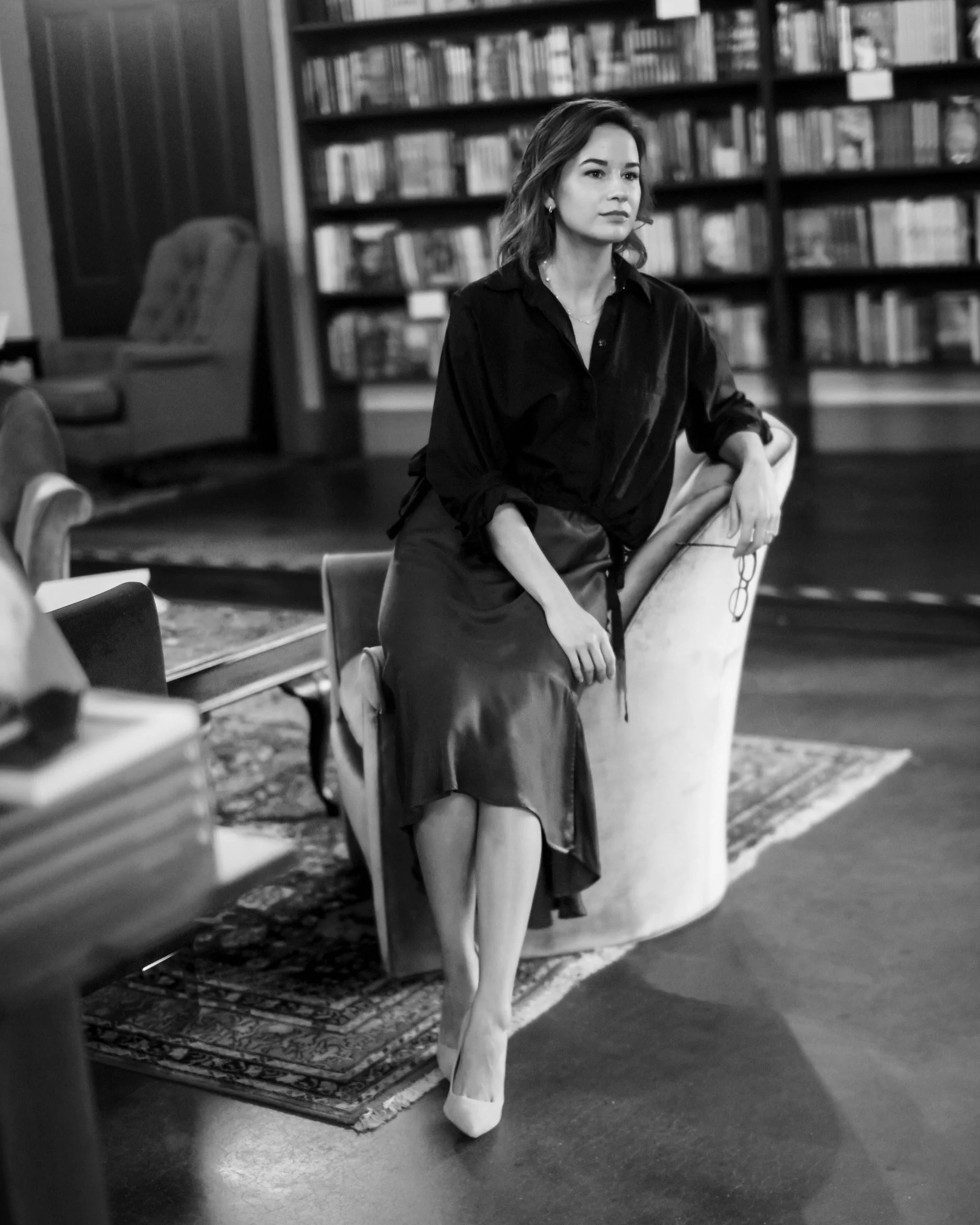 A woman with shoulder-length hair sitting on a chair in a library, with bookshelves in the background, dressed in a dark blouse and a long skirt, holding eyeglasses in her hand.