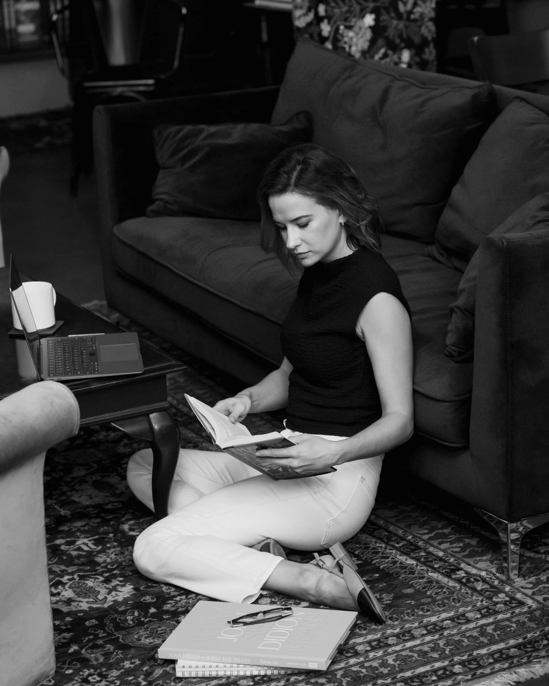 A woman with short hair and in a black top and white pants sits on a patterned rug on the floor, reading a magazine. There are books, glasses, and a laptop on a small table beside her, and a sofa with cushions behind her.