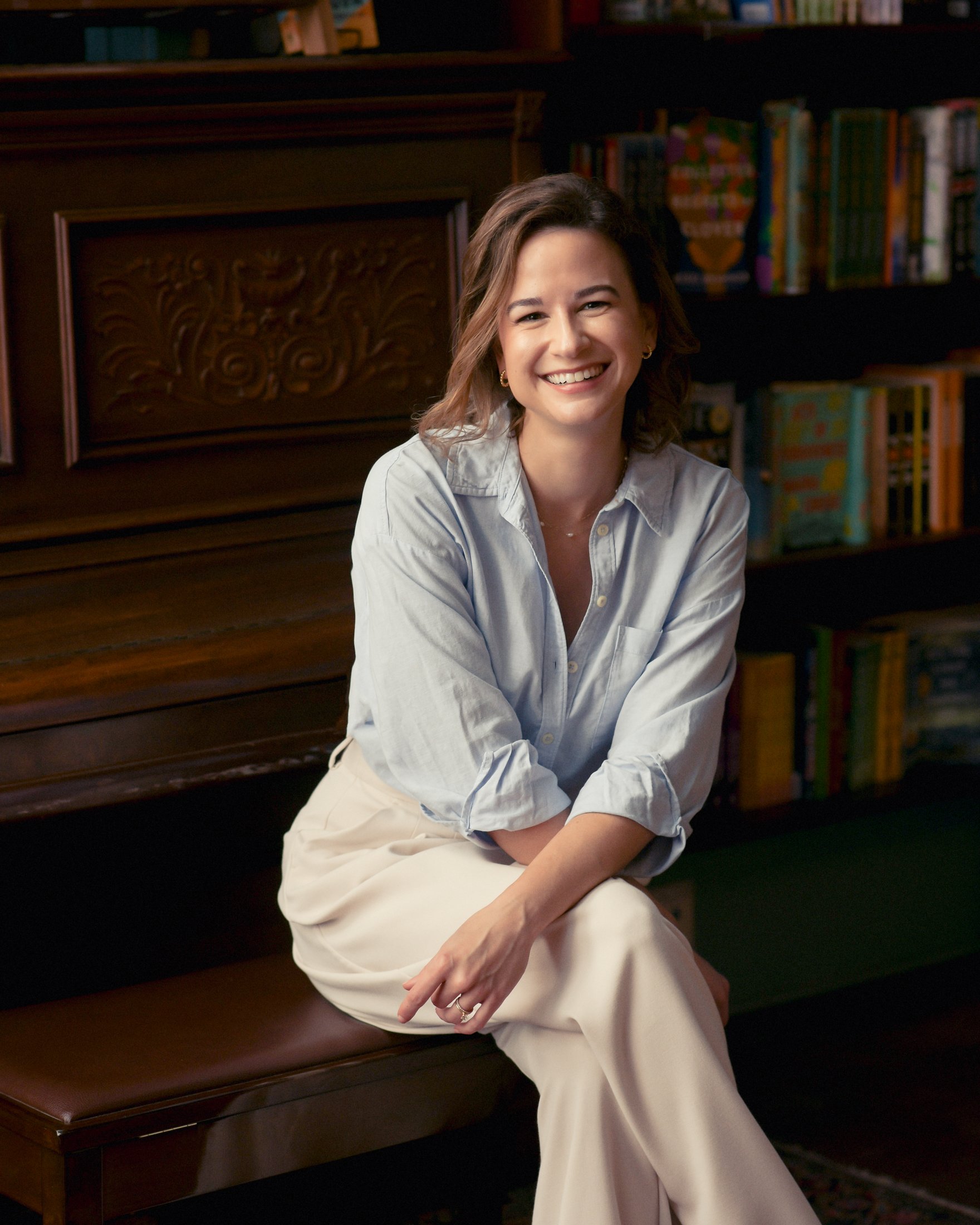 A woman with shoulder-length brown hair, wearing a light blue button-up shirt and cream-colored pants, sitting on a wooden bench, smiling with a background of a dark wood bookshelf filled with colorful books.