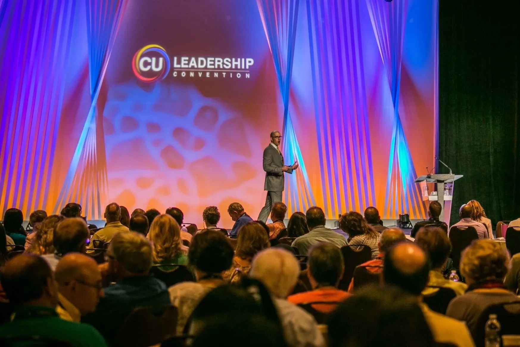 A speaker on stage at a CU Leadership Convention addressing an audience, with a large purple and orange backdrop displaying the CU Leadership logo.