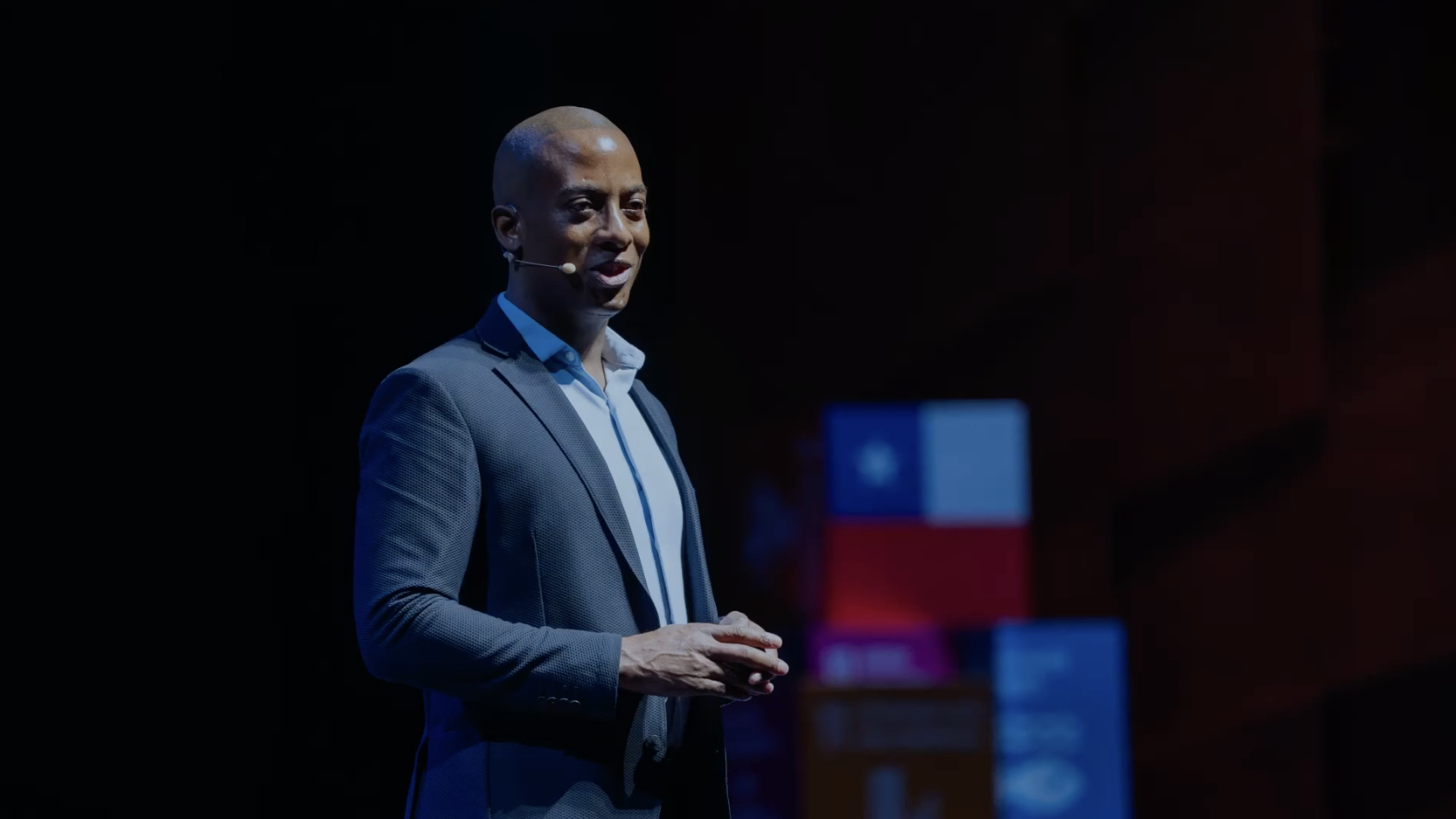 A man in a blue suit giving a presentation on stage with a microphone headset, standing in front of a dark background with illuminated boxes in the distance.
