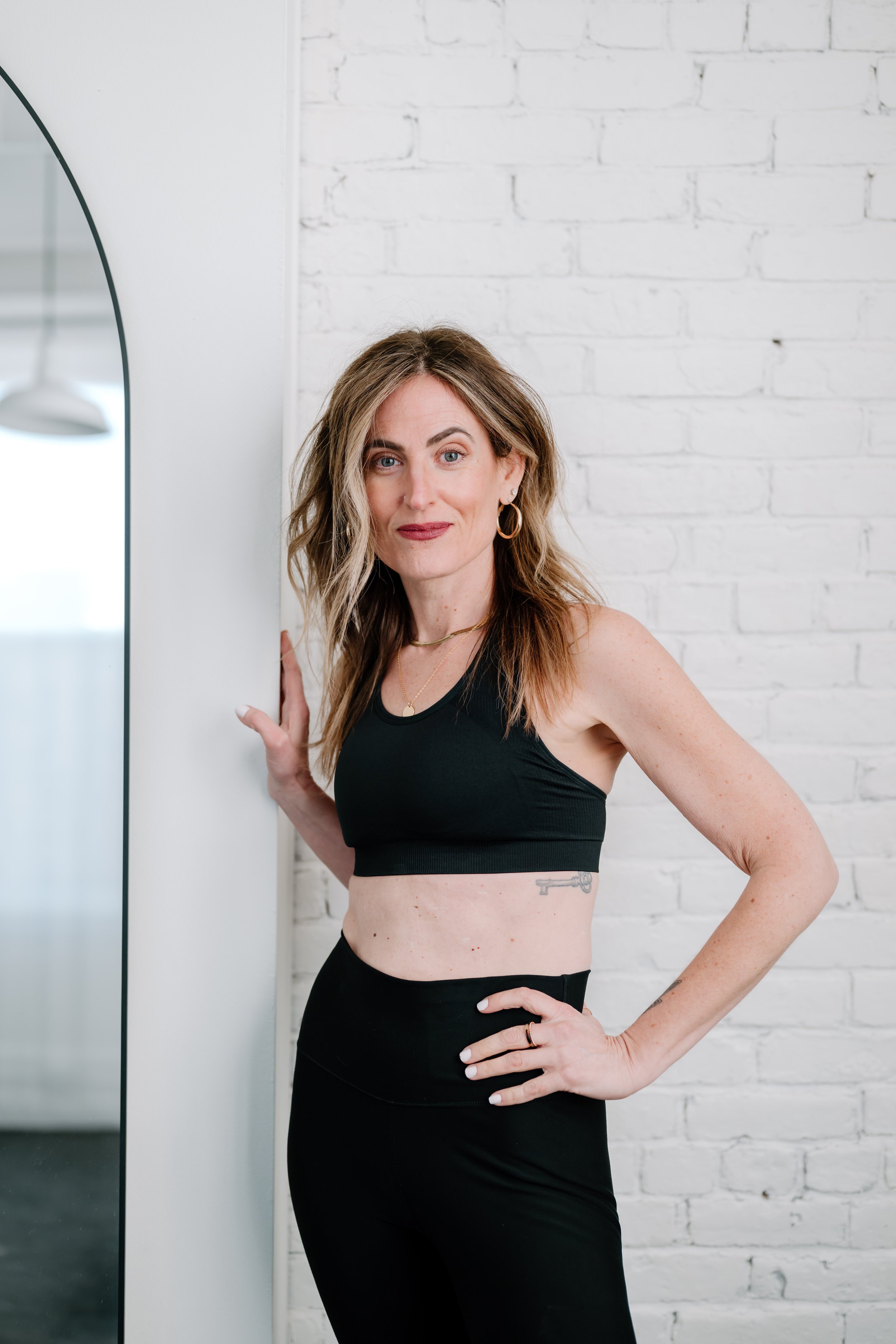 A woman smiling and flexing her right arm while holding a dumbbell in her left hand. She is wearing a black graphic t-shirt the word "Queen" and black leggings, standing against a white wall.
