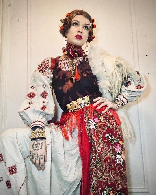 Woman dressed in traditional Eastern European or Balkan folk attire, wearing embroidered blouse, a red and gold embroidered skirt, and various jewelry, with hair styled in rollers and makeup.