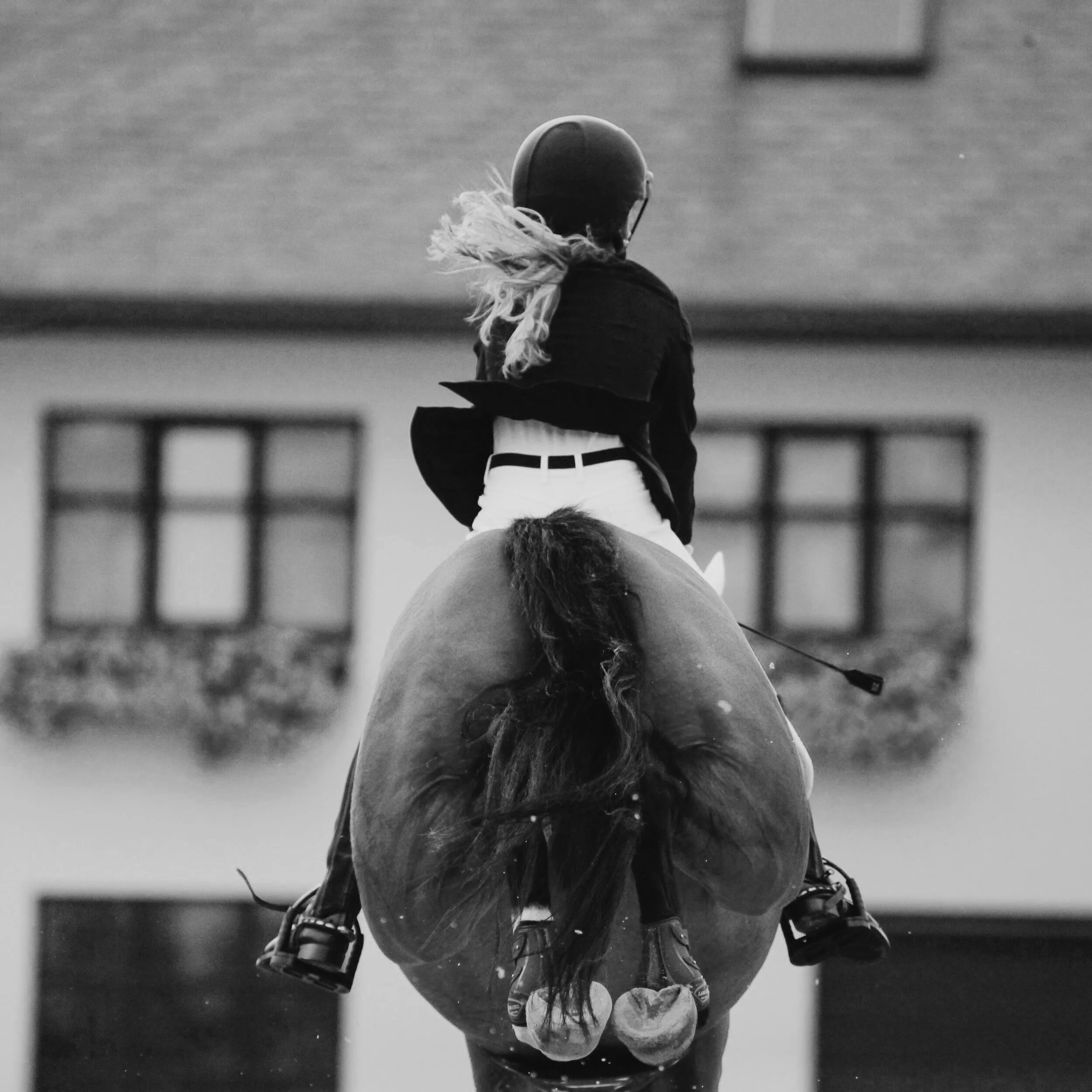 A woman riding a horse, viewed from behind, in black and white. She is wearing a helmet, a dark jacket, and white pants, with her hair flying in the wind. The house and windows are blurred in the background.