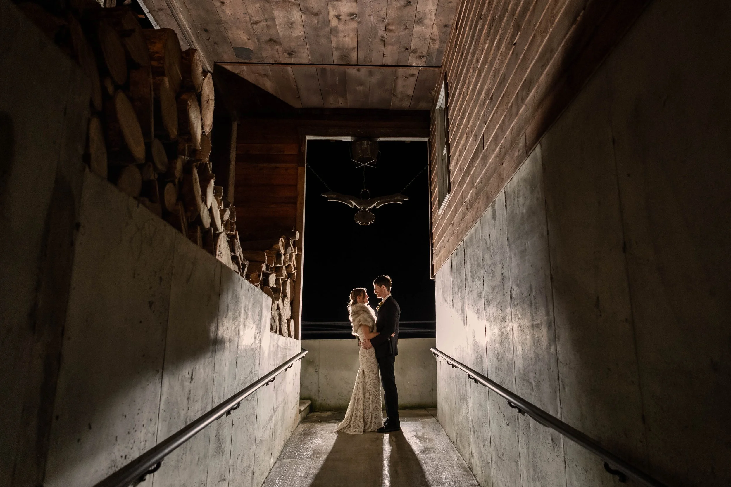 A couple in wedding attire standing close together on an outdoor walkway at night, framed by a rustic building and woodpile, with a dark night sky and a decorative outdoor chandelier overhead.