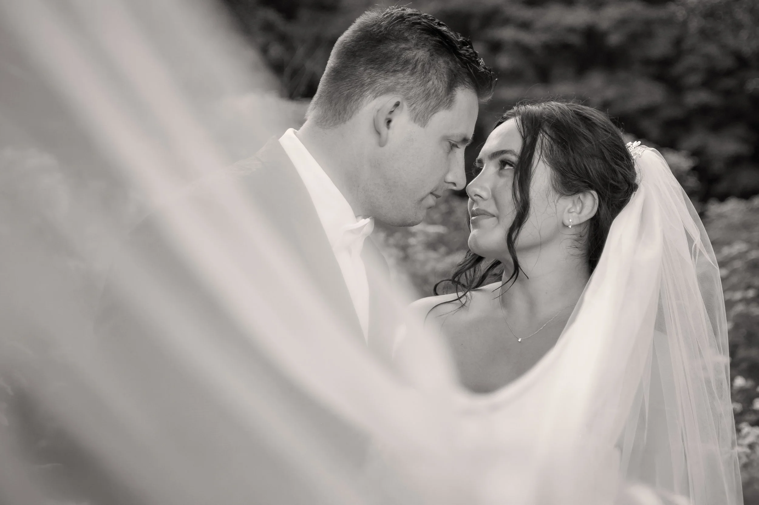 A black and white photograph of a bride and groom close together, gazing into each other's eyes, with the bride wearing a veil and wedding dress, and the groom in a suit.