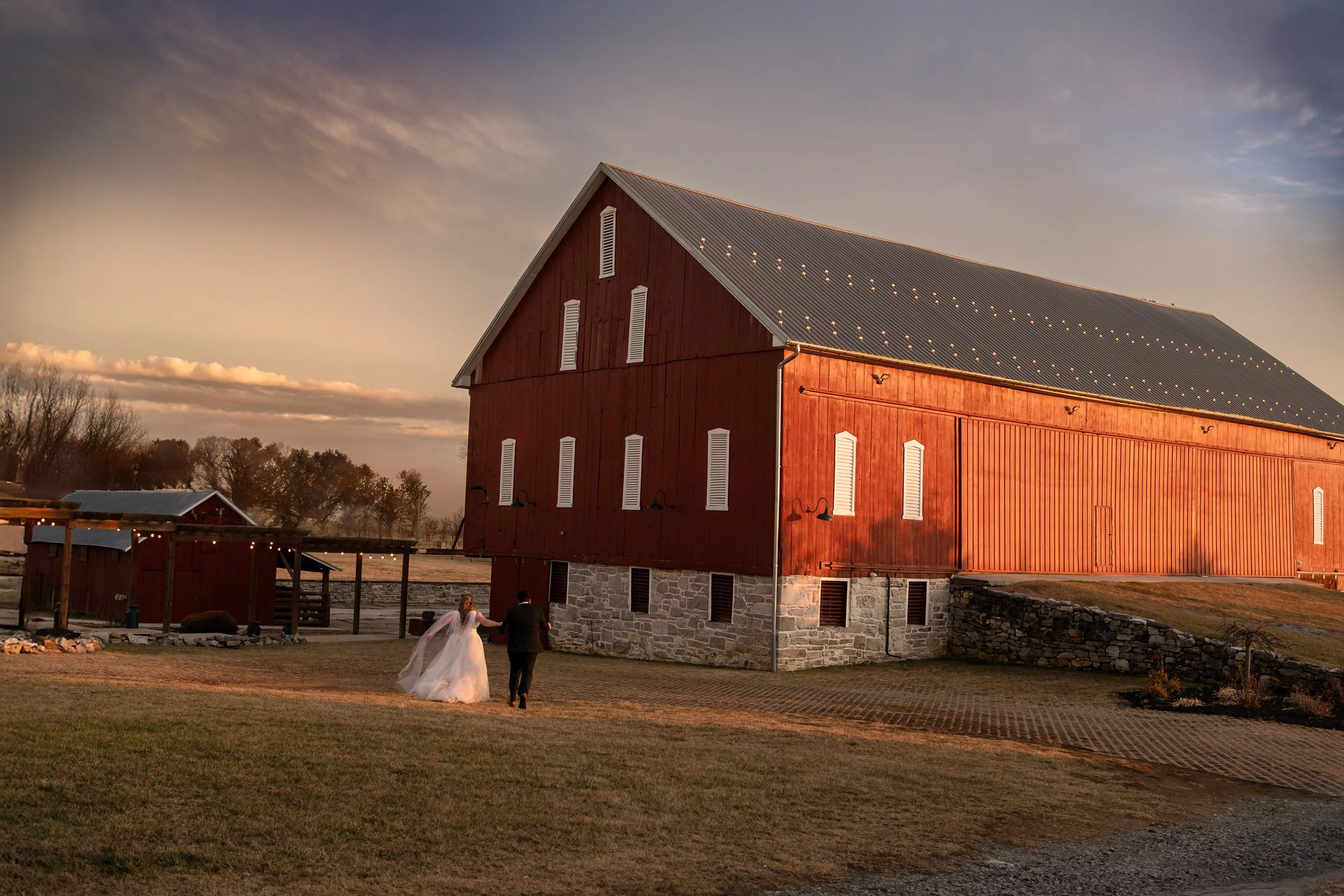 A bride and groom walking together outside a large red barn at sunset.