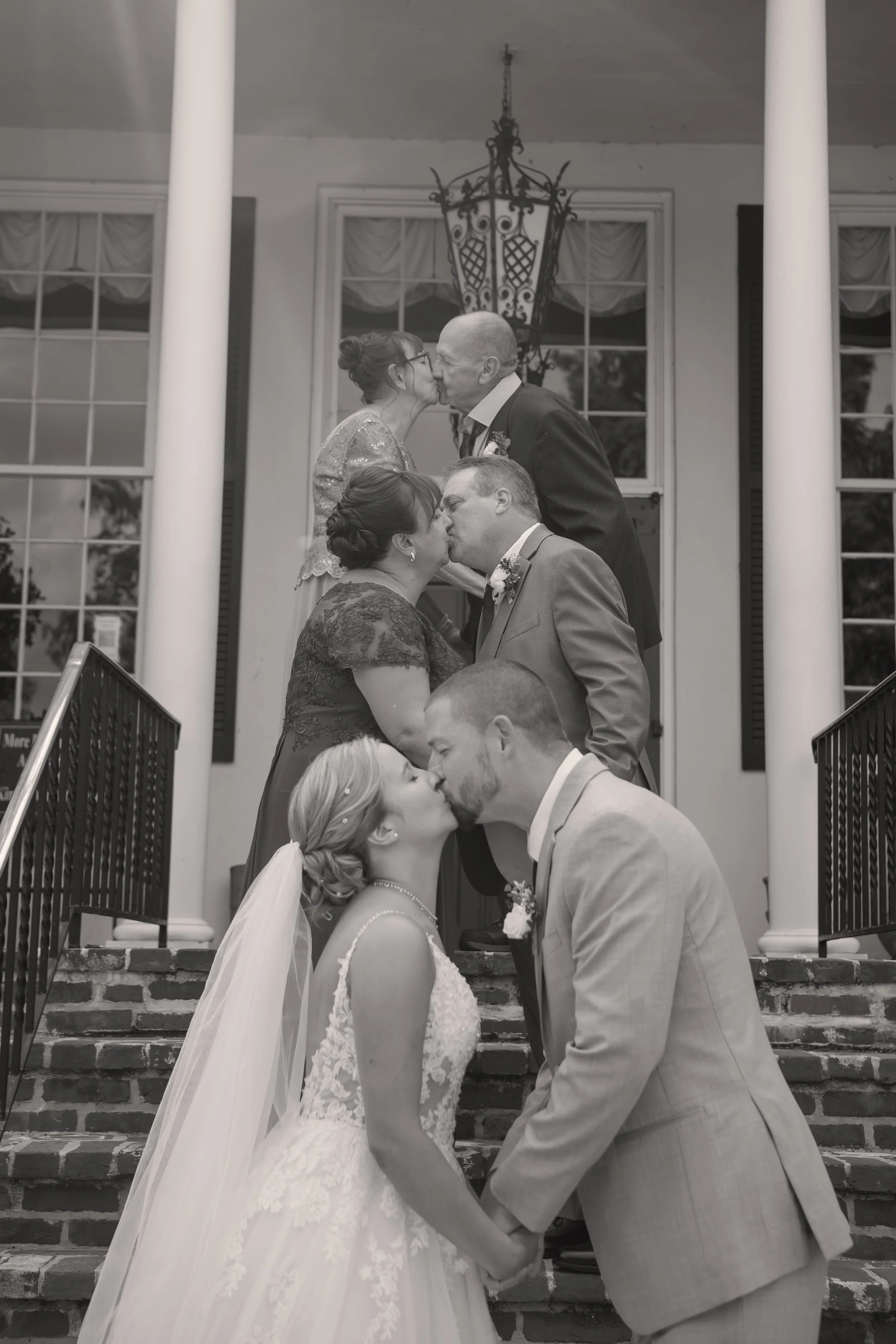 Six couples, arranged vertically, kiss on a staircase outside a building with large windows and a hanging lantern.