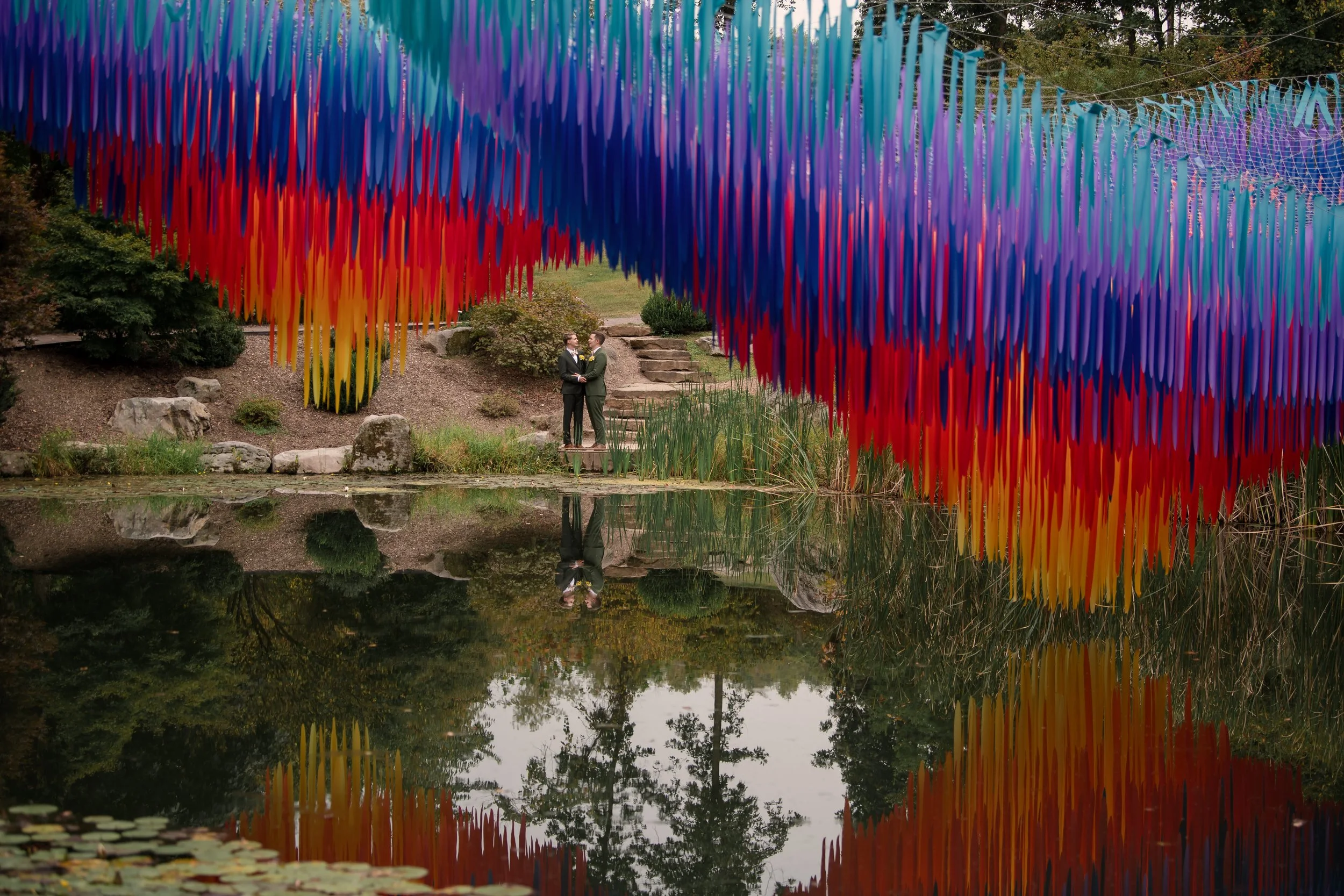 A rainbow-colored art installation hanging above a pond with two people in formal attire standing on the opposite side, reflected in the water surrounded by greenery and trees.