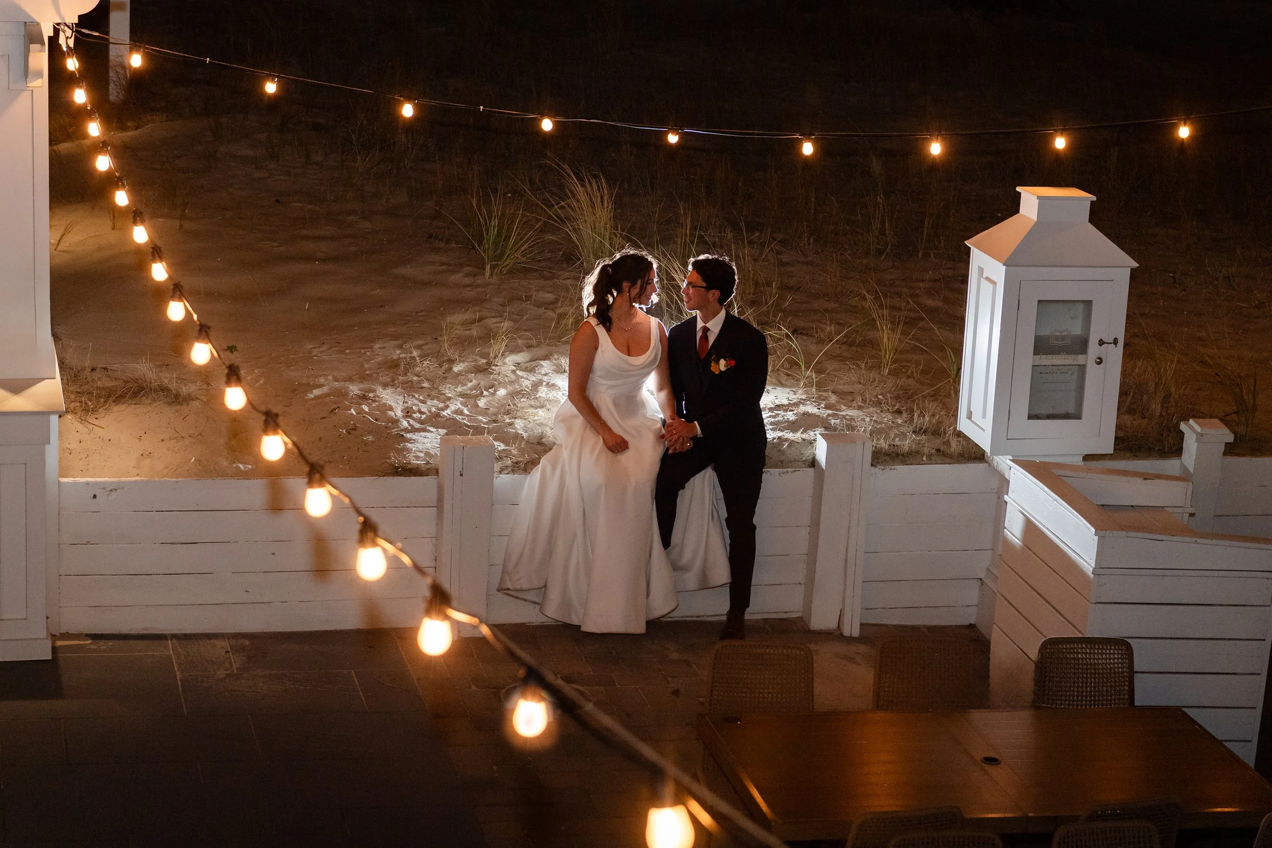 A bride and groom sitting on a white fence at night, holding hands, with string lights overhead and a beach background.