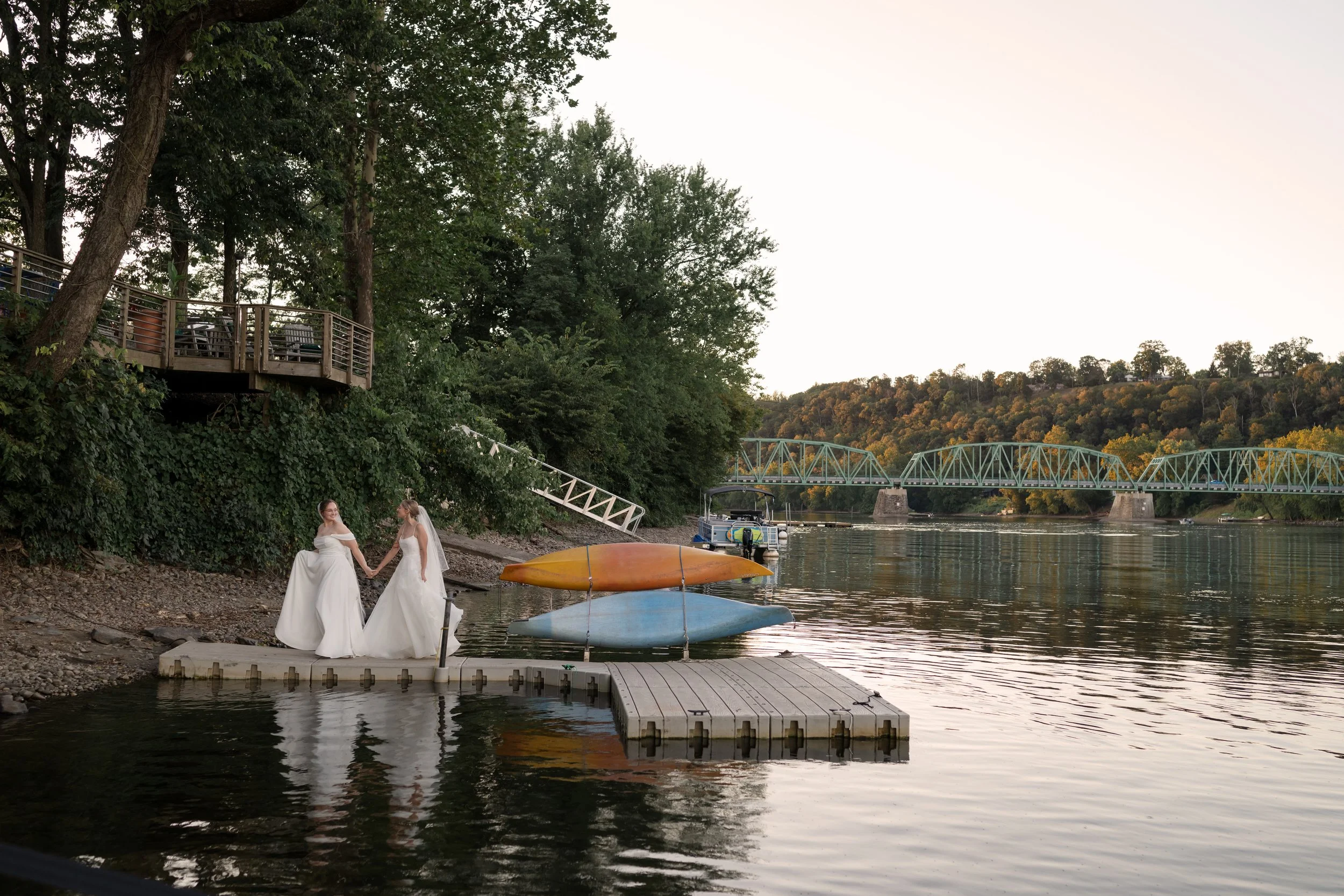 Two women in wedding dresses holding hands on a dock by a river, with kayaks and a boat in the background, trees on the shoreline, and a bridge over the water during sunset.