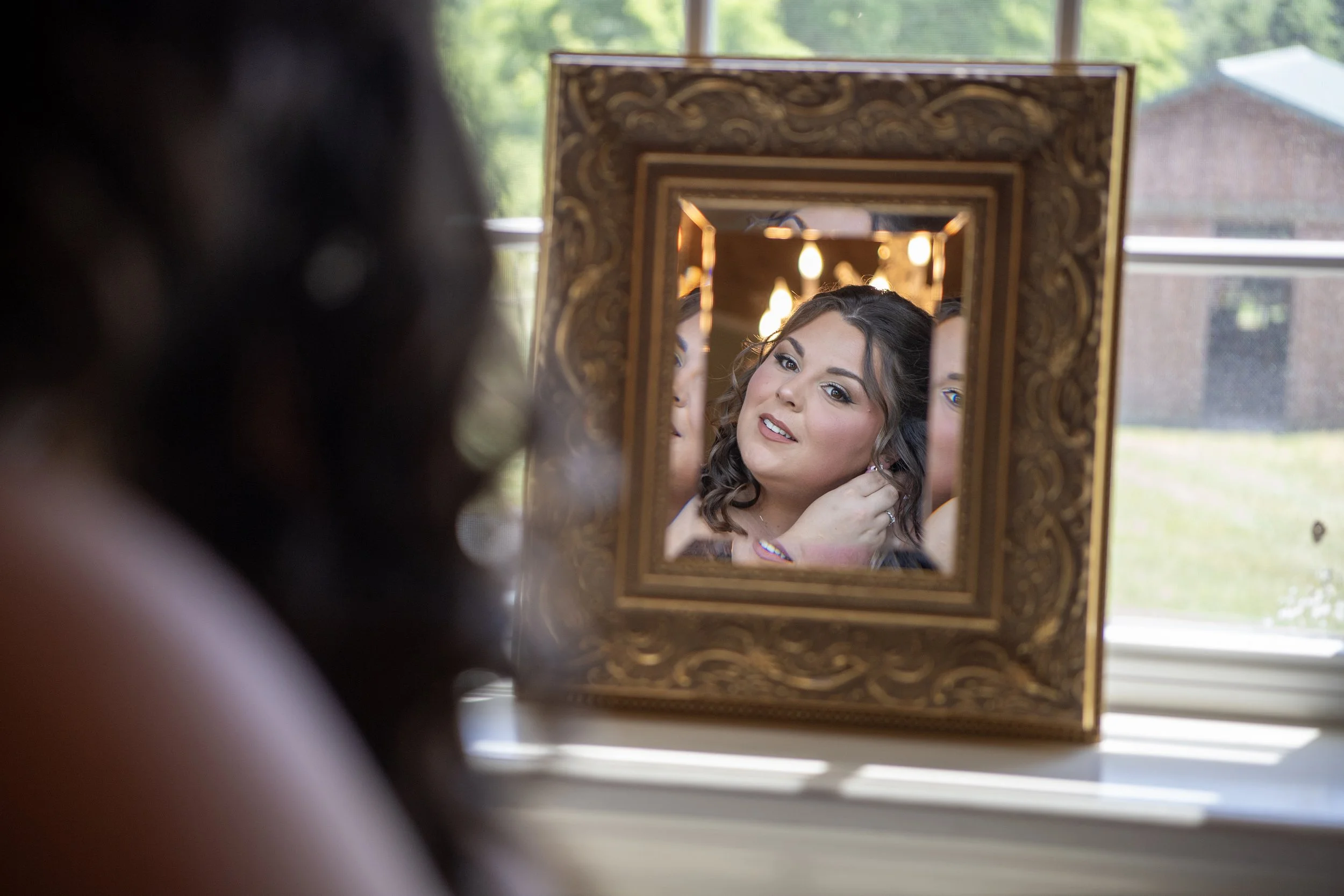 A woman looking into a decorative gold-framed mirror, adjusting her earring, with her reflection showing her makeup and hairstyle, in a well-lit room with large windows.