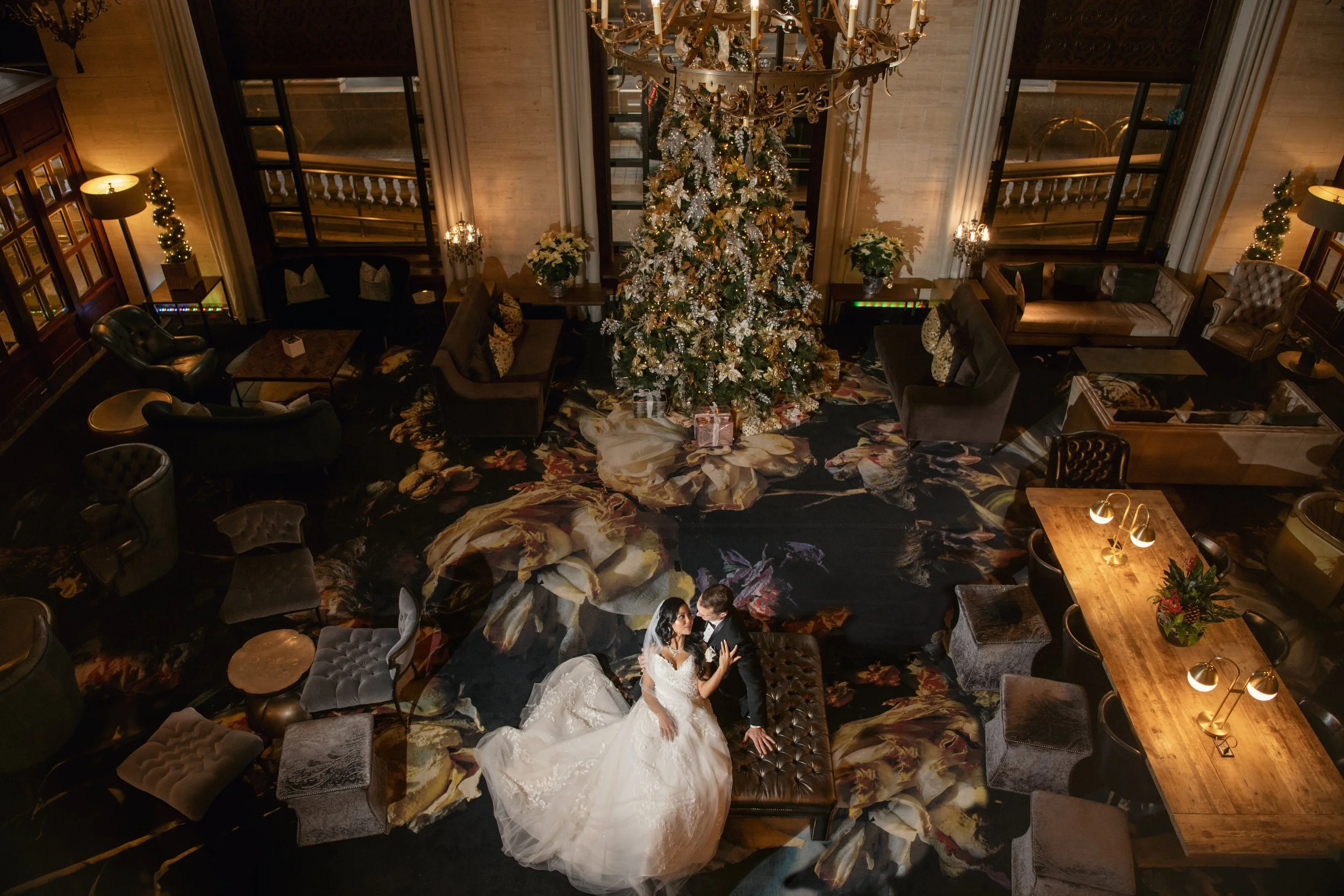 A bride and groom dancing on a tufted ottoman in a luxurious, dimly-lit hotel lobby decorated for Christmas with a large decorated Christmas tree, presents underneath, a floral rug, and elegant furniture, including couches and armchairs, surrounding 