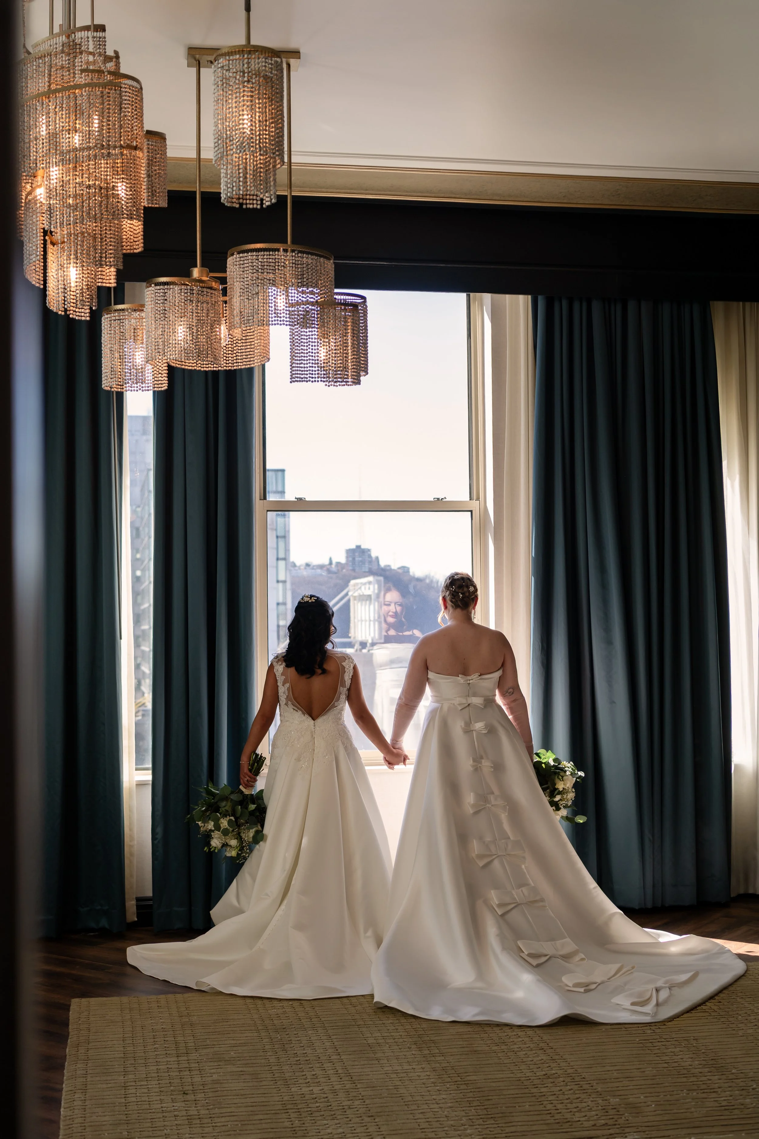 Two brides in wedding dresses holding hands and holding bouquets, standing in front of a large window with cityscape view, in a decorated room with hanging chandeliers and curtains.