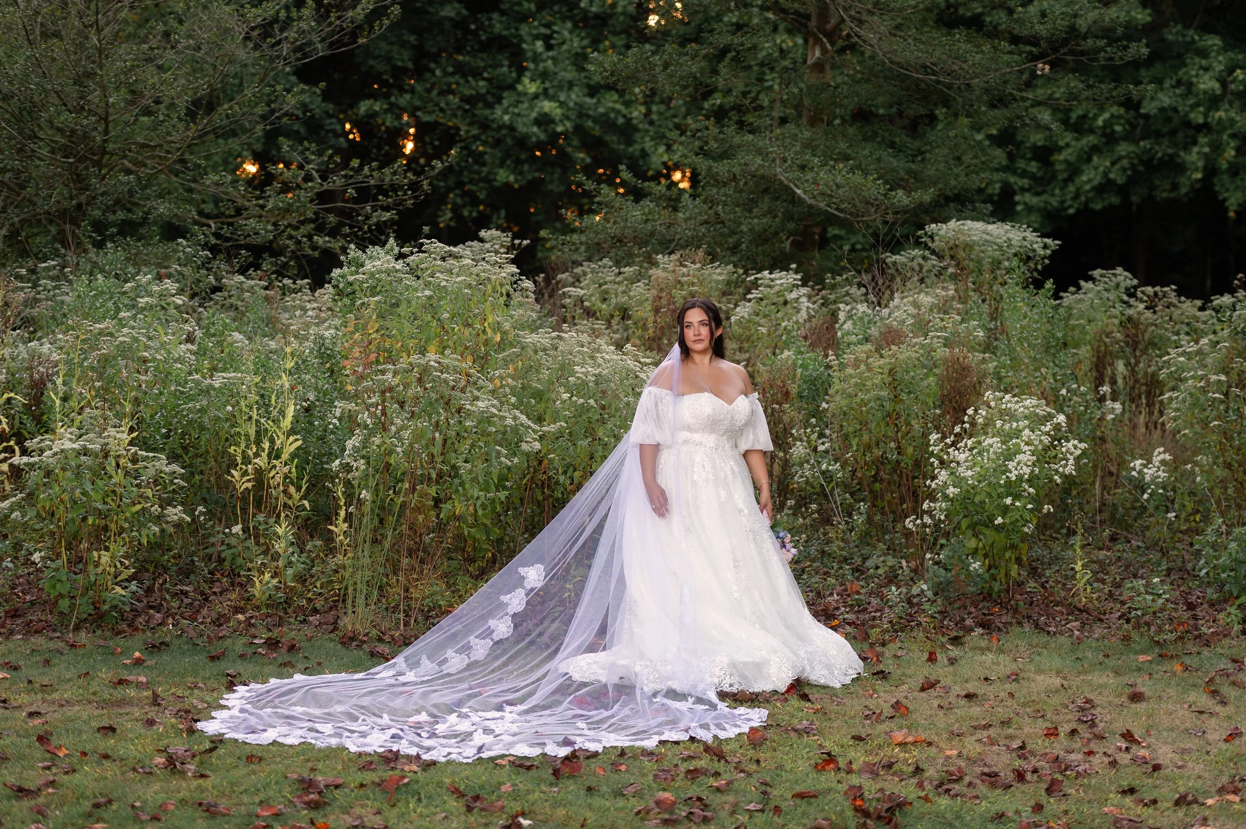 A woman in a white wedding gown with a long train and veil stands in a garden with white flowers and green foliage.