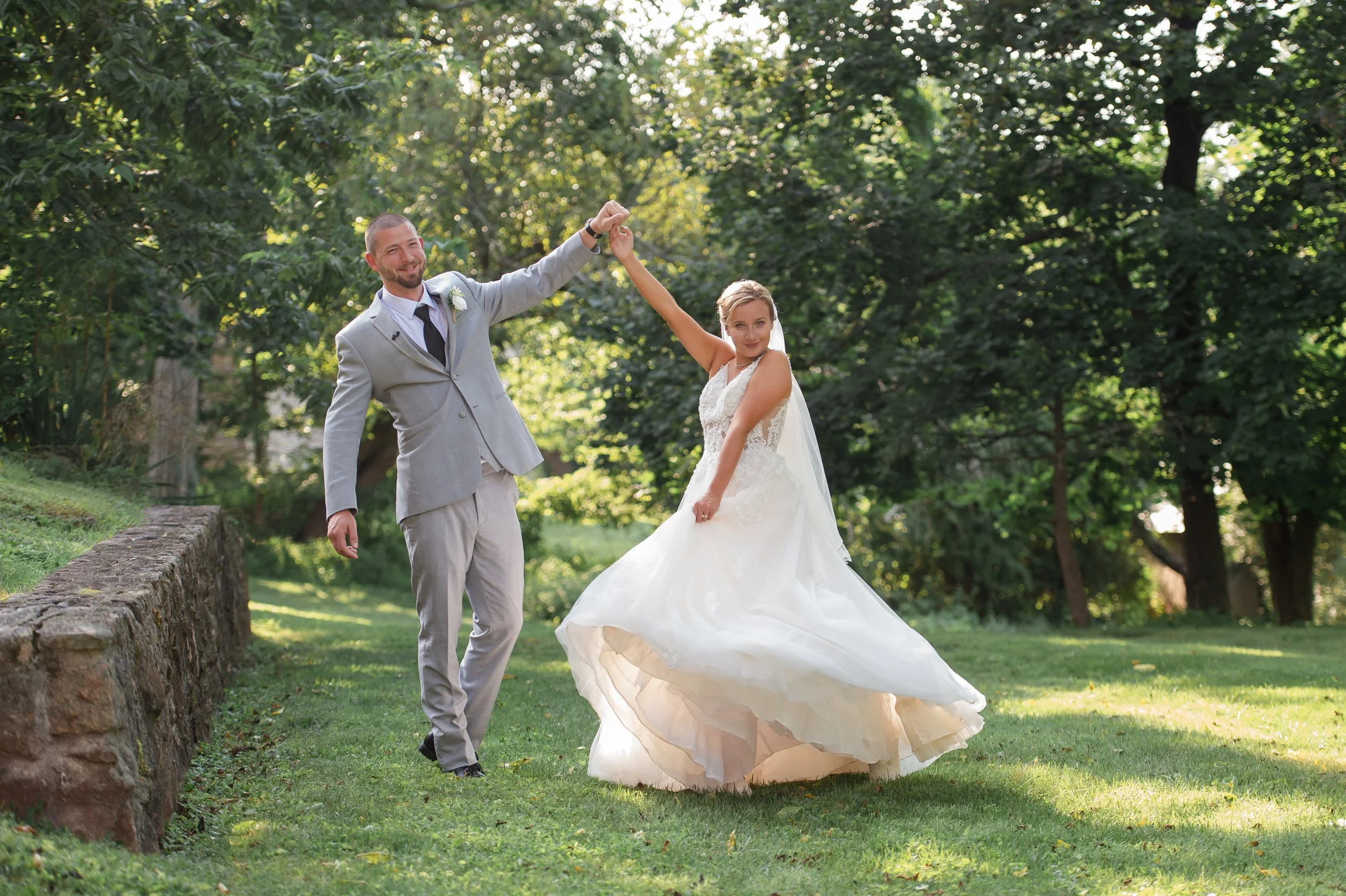 A bride and groom dancing outdoors in a lush green park. The groom, dressed in a light gray suit, holds the bride's hand as she twirls in her white wedding gown. Sunlight filters through the trees.