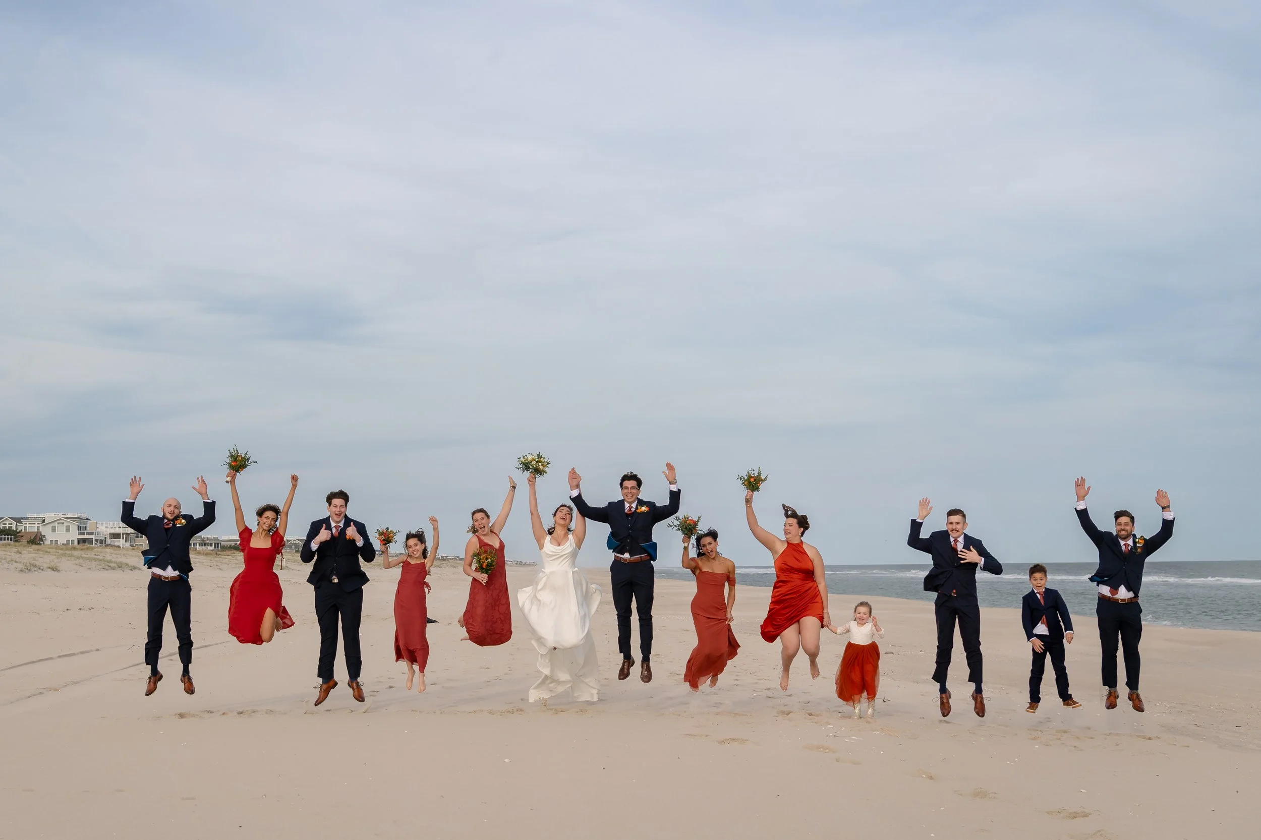 A wedding party of twelve jumping on a beach with the ocean in the background, some holding bouquets and others holding hands.
