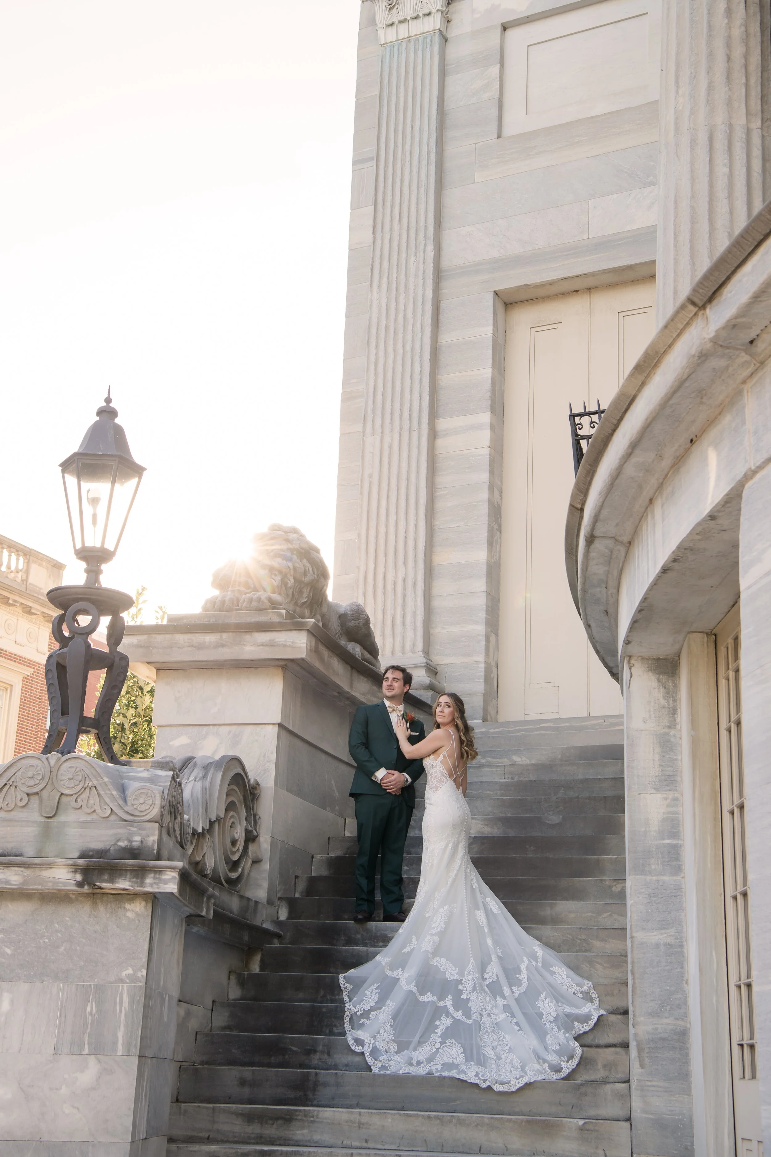 Bride and groom standing on stone stairs outside a historic building, with lion sculptures and a street lamp, during sunset.