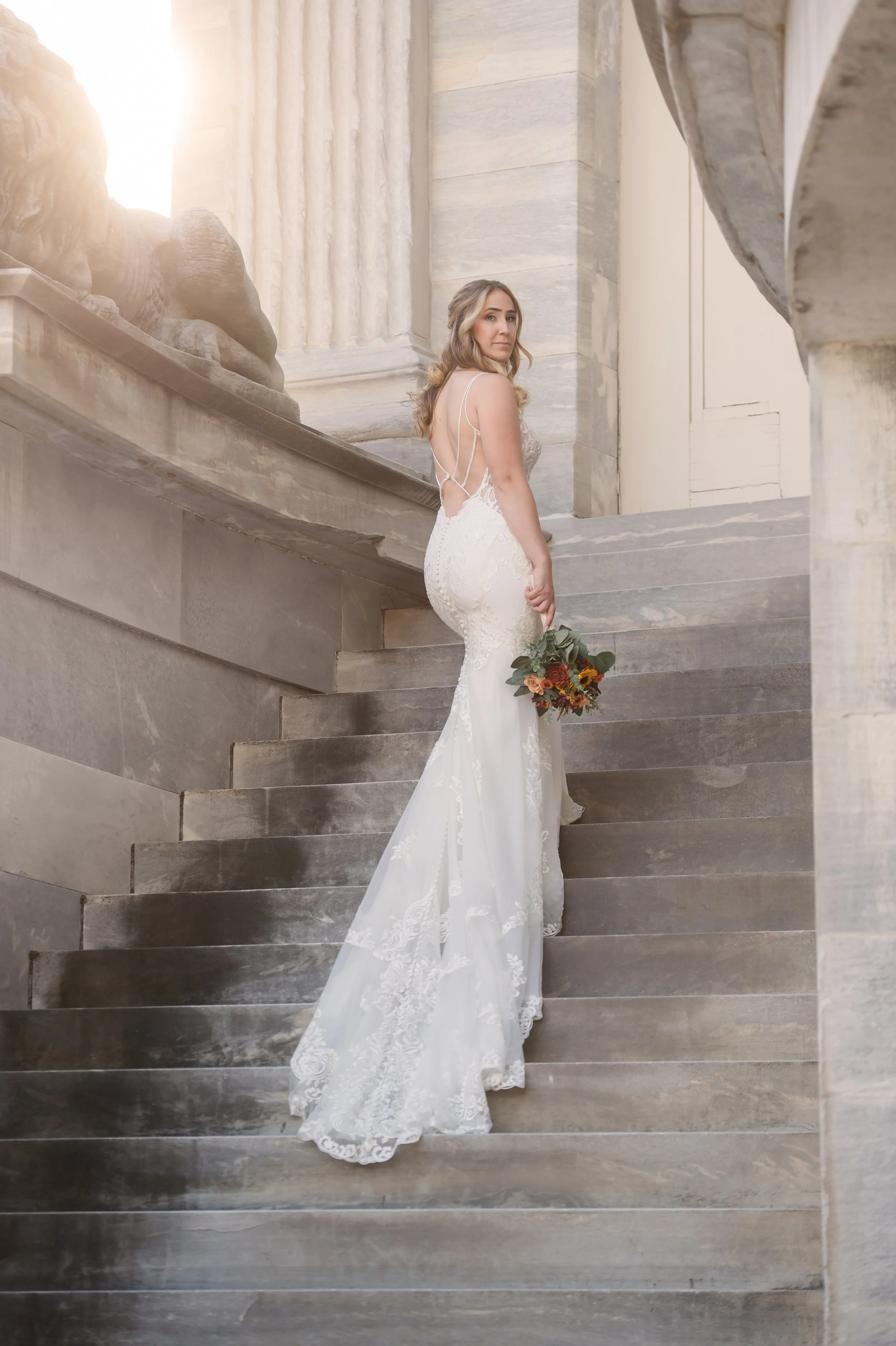 A bride in a white lace wedding gown holding a bouquet, standing on a grand stone staircase inside a building with classical architecture.