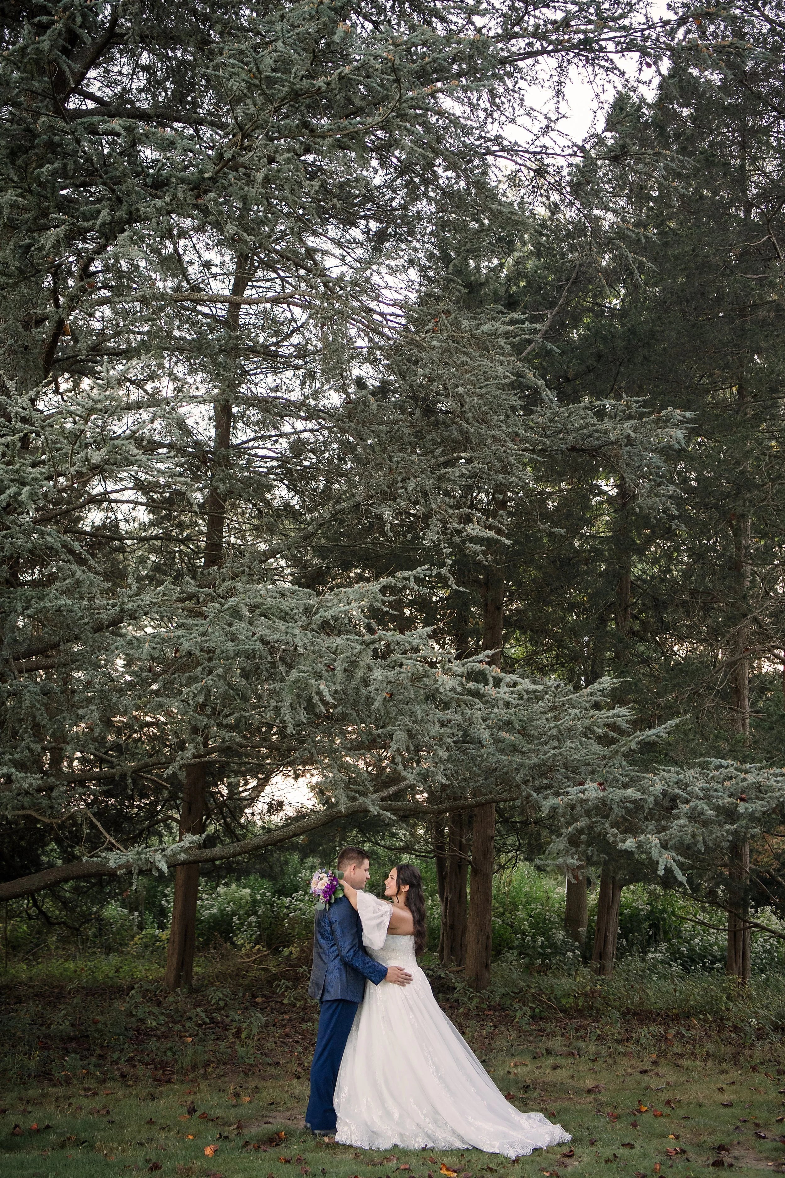 A bride and groom standing close together on grass in front of tall trees. The bride is wearing a white wedding gown, and the groom is dressed in a blue suit. The bride is holding a bouquet of purple and white flowers, and they are gazing at each oth
