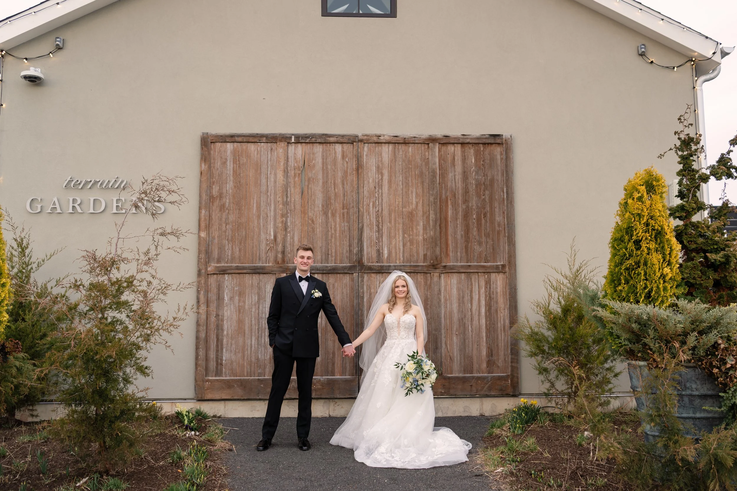 A bride and groom standing outside a rustic wooden barn on their wedding day. The bride wears a white strapless wedding gown with a veil and holds a bouquet. The groom wears a black tuxedo with a bow tie and white boutonniere. They are holding hands,