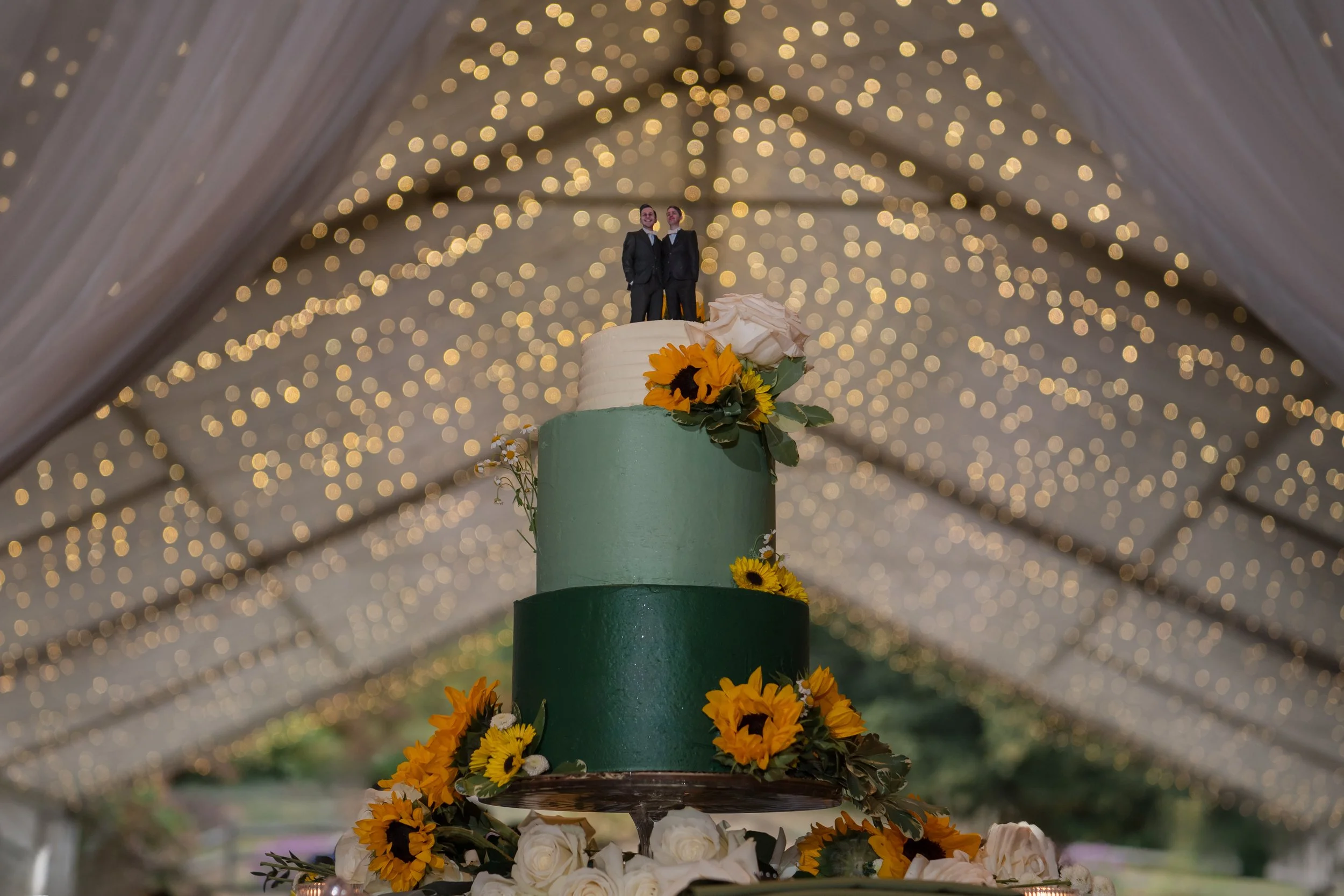 A multi-tiered wedding cake decorated with sunflowers and white roses, topped with cake toppers of two grooms in tuxedos, placed under a tent with string lights.