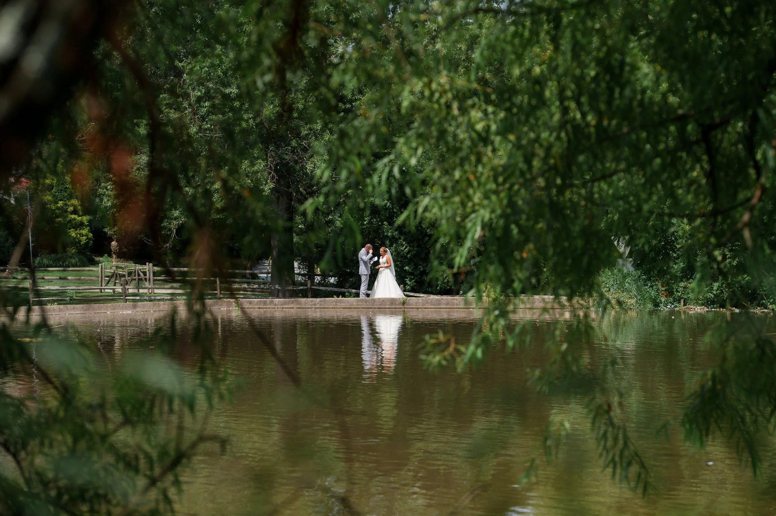 A bride and groom standing on a narrow pathway by a body of water, framed by overhanging tree branches, with greenery and trees in the background.