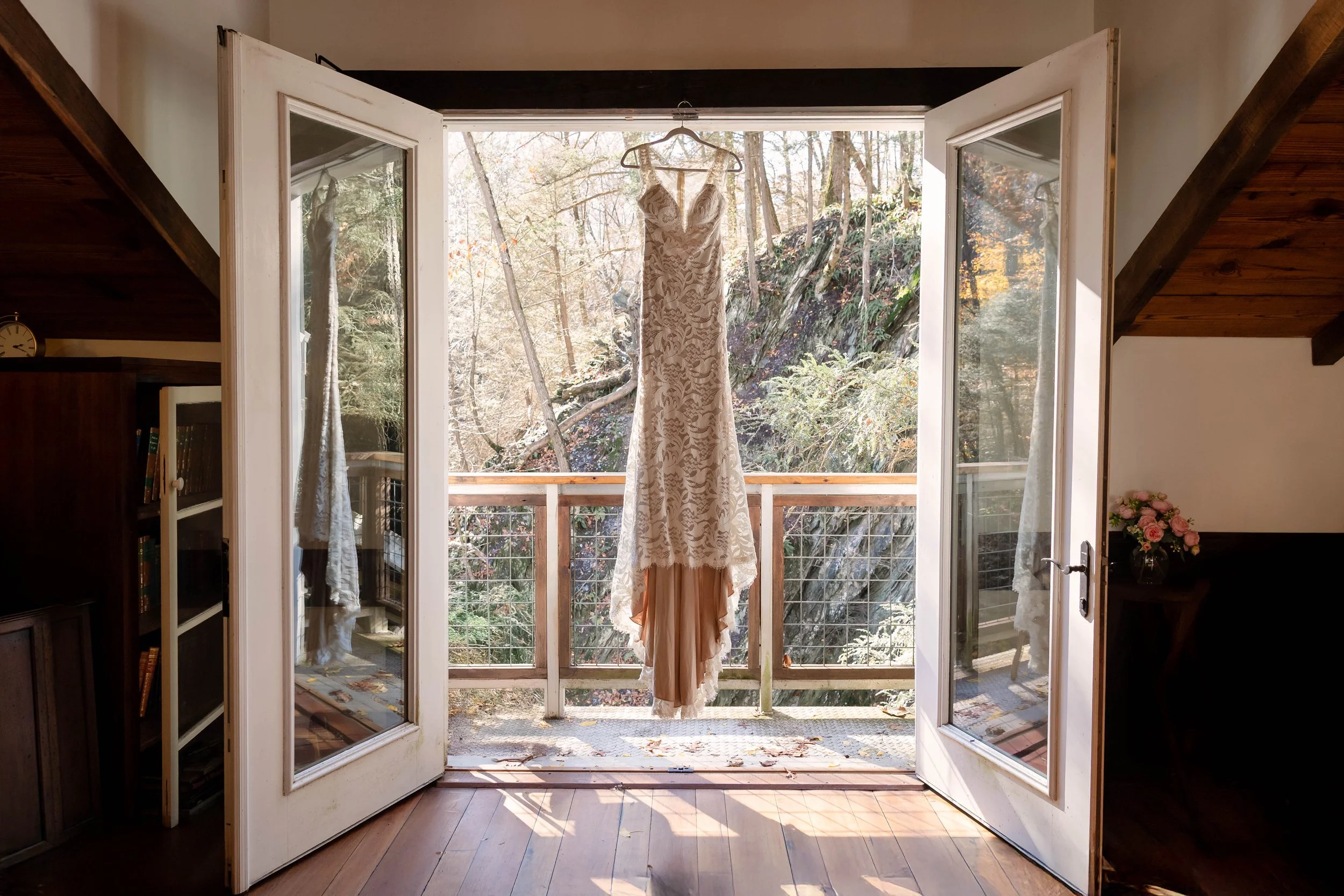 A white lace wedding dress hanging on a hanger outside a glass door, with a wooded area in the background.