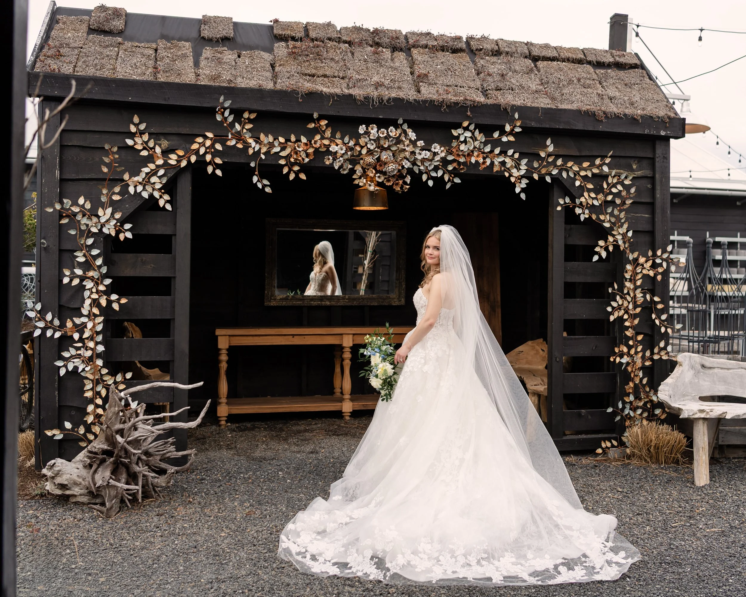 A bride in a wedding dress holding a bouquet, standing outside in front of a black wooden structure decorated with metal leaf accents, with her reflection visible in a mirror behind her.