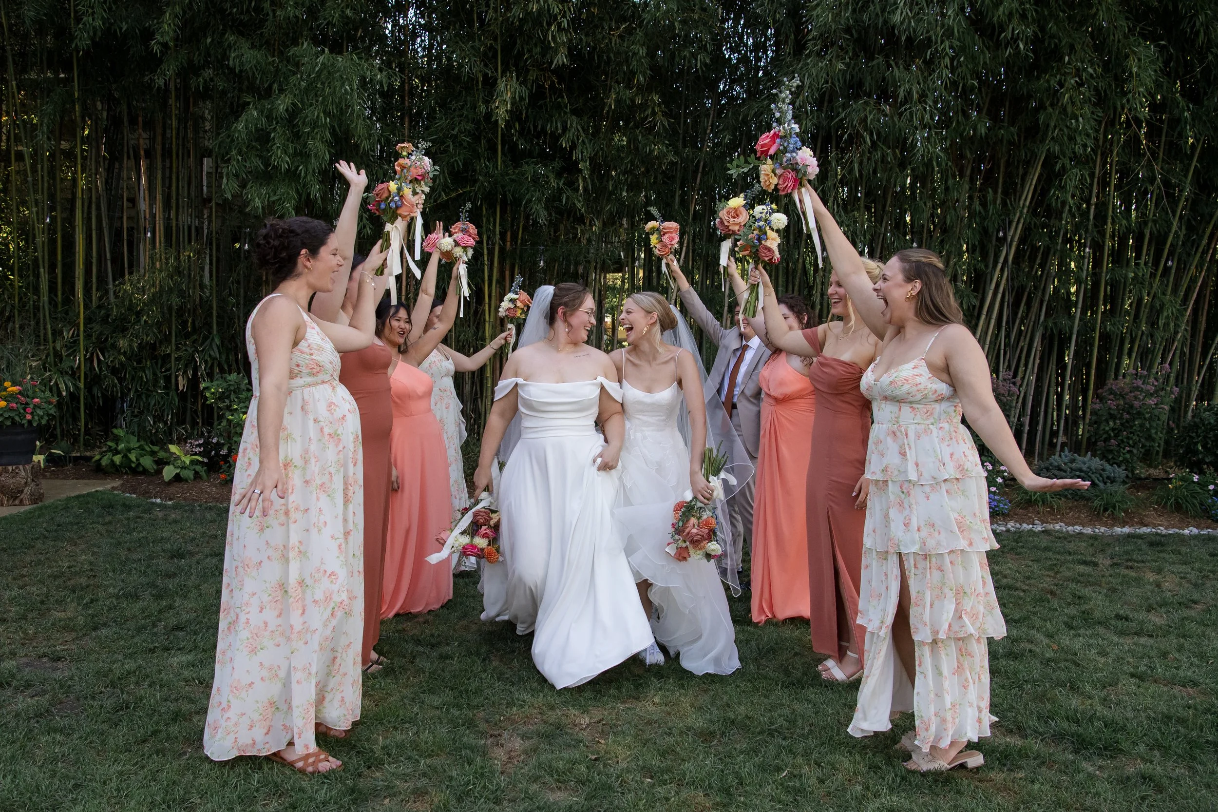Two brides in white wedding dresses walking together, surrounded by friends raising bouquets and celebrating outdoors with greenery and bamboo in the background.