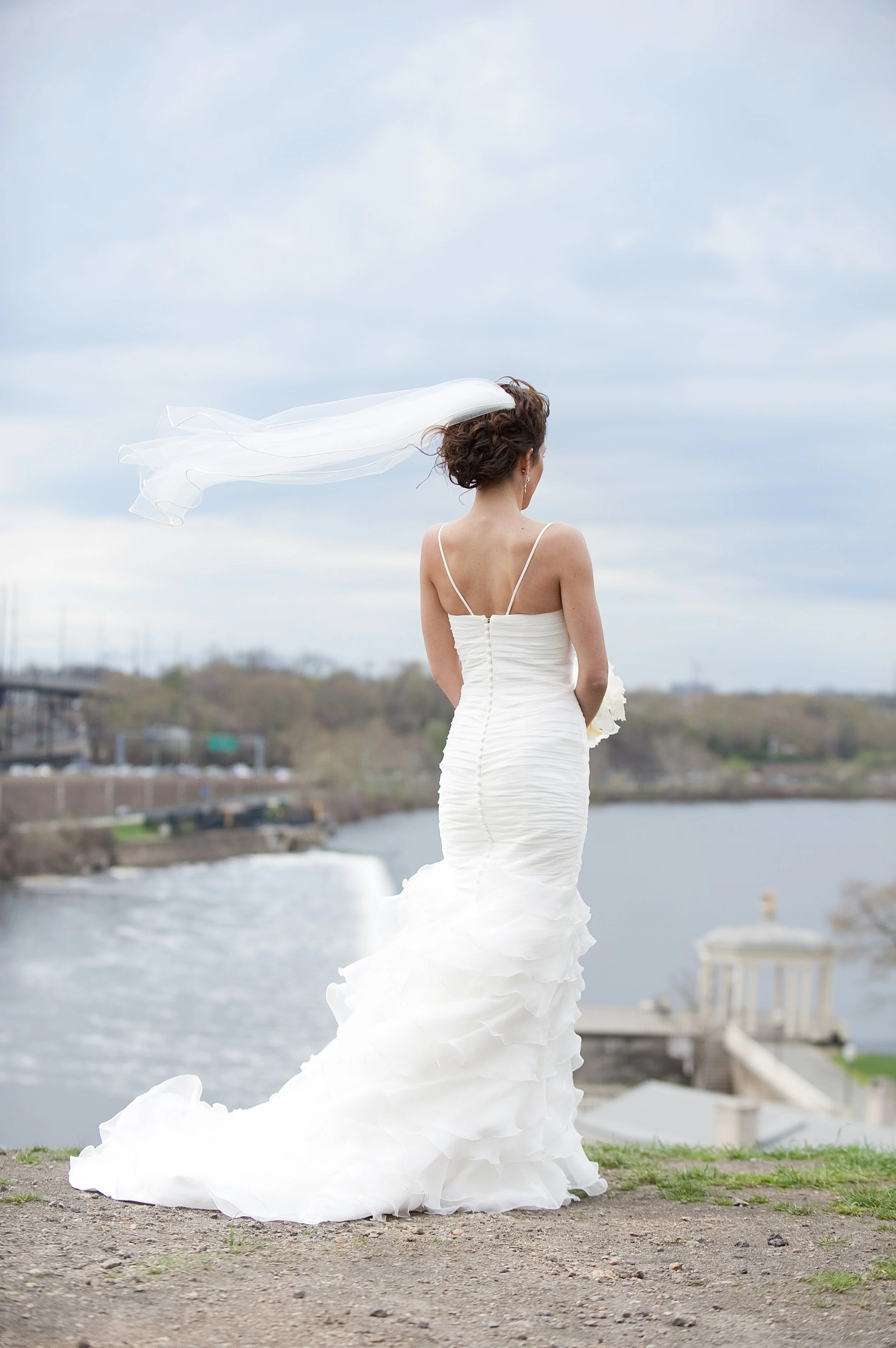 A bride standing outdoors near a body of water, wearing a white wedding dress with a long, ruffled train and a veil blowing in the wind, with a cloudy sky in the background.