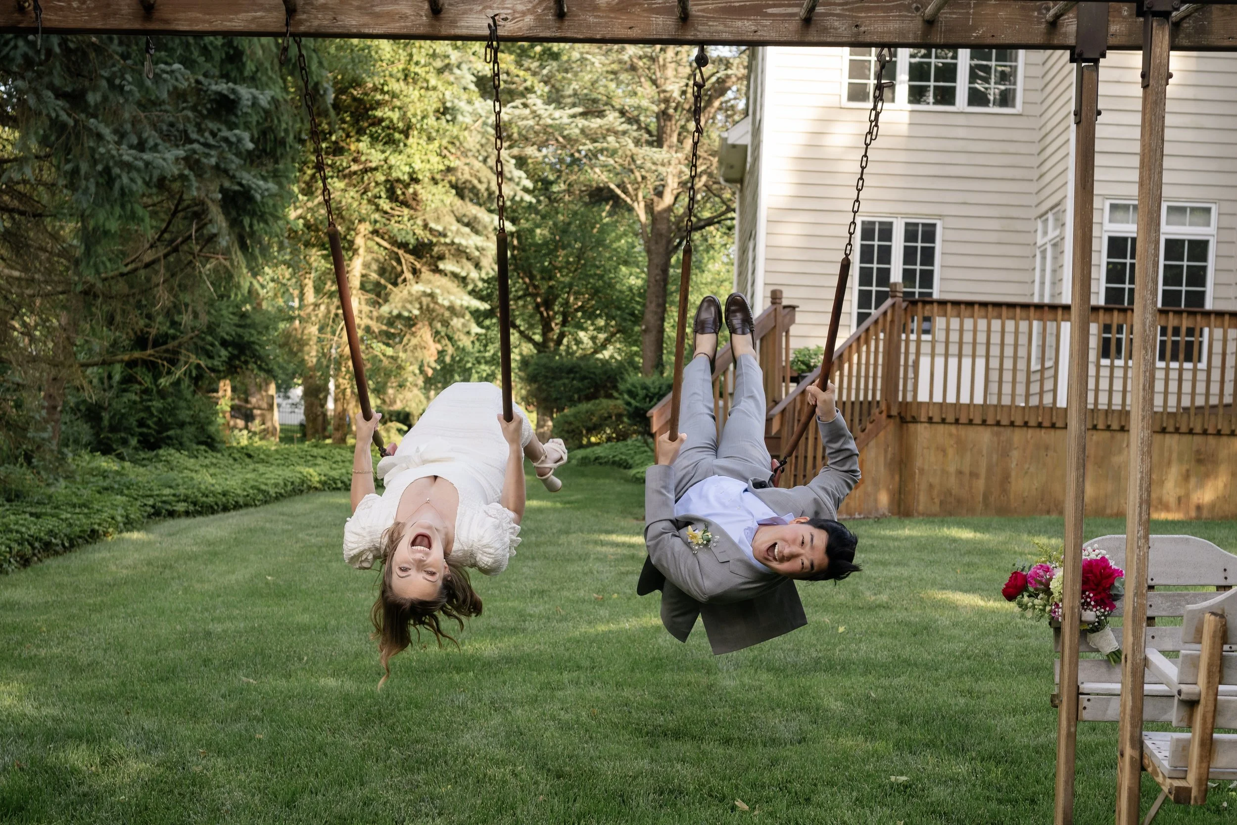 Two people swinging on a swing set in a backyard, both smiling and looking happy. The woman is wearing a white dress, and the man is dressed in a gray suit. There is a house with wooden stairs and a deck in the background, along with green trees and 
