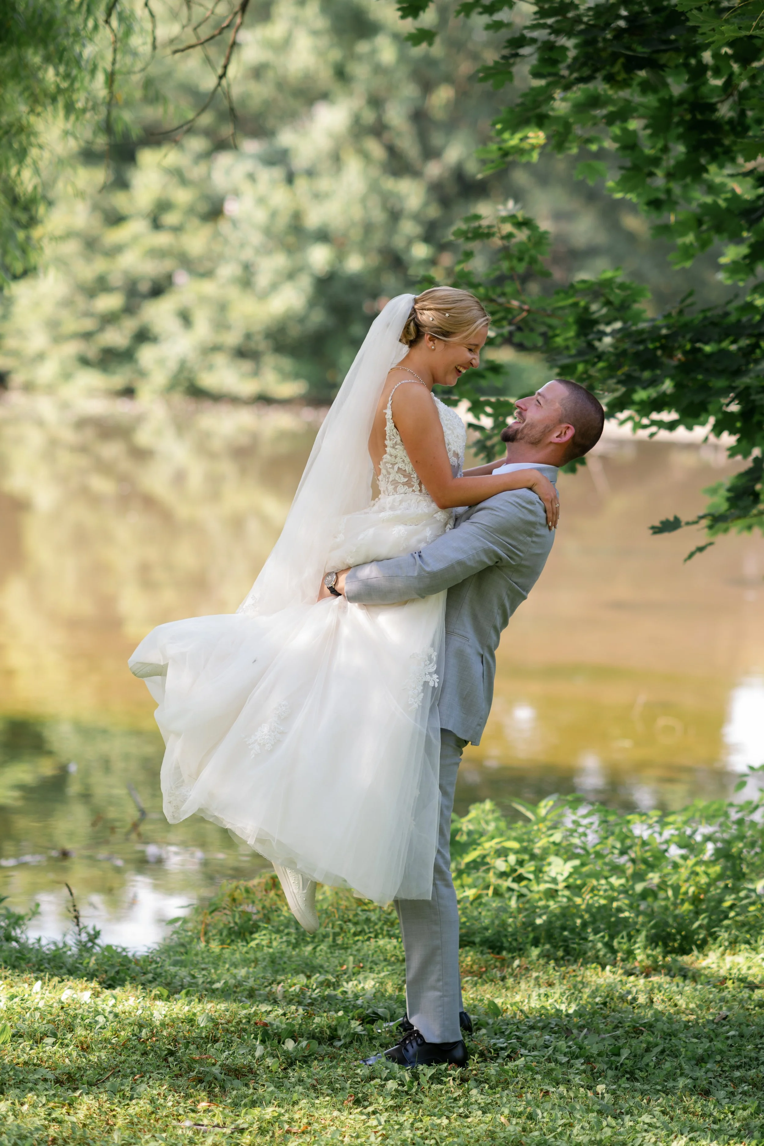 A happy bride and groom are outdoors near a lake, with the groom lifting the bride in his arms. The bride is smiling and wearing a white wedding gown with a veil, and the groom is smiling back, dressed in a light gray suit. Surrounded by green trees 
