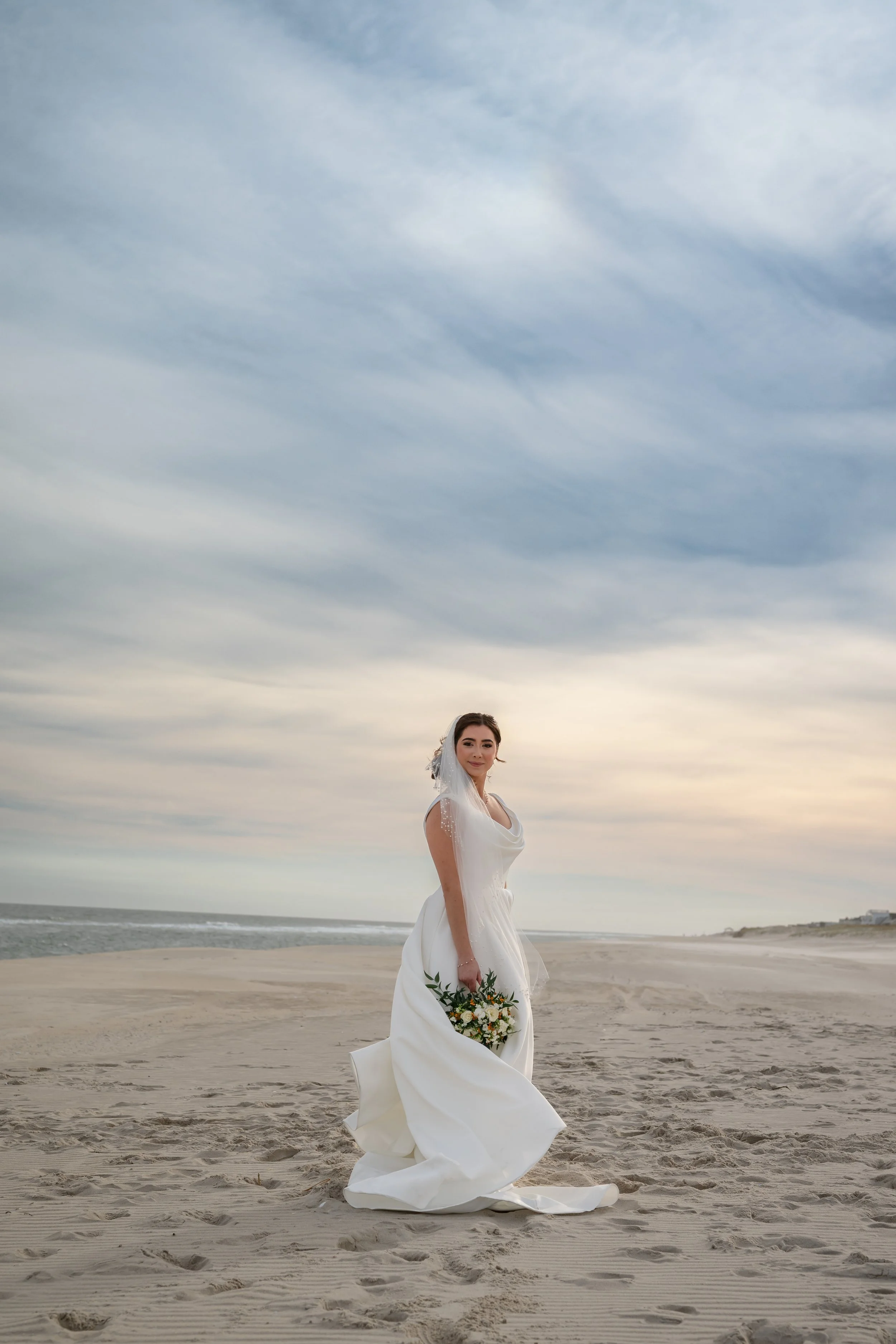 A bride in a white wedding dress holding a bouquet of flowers on a sandy beach during sunset.