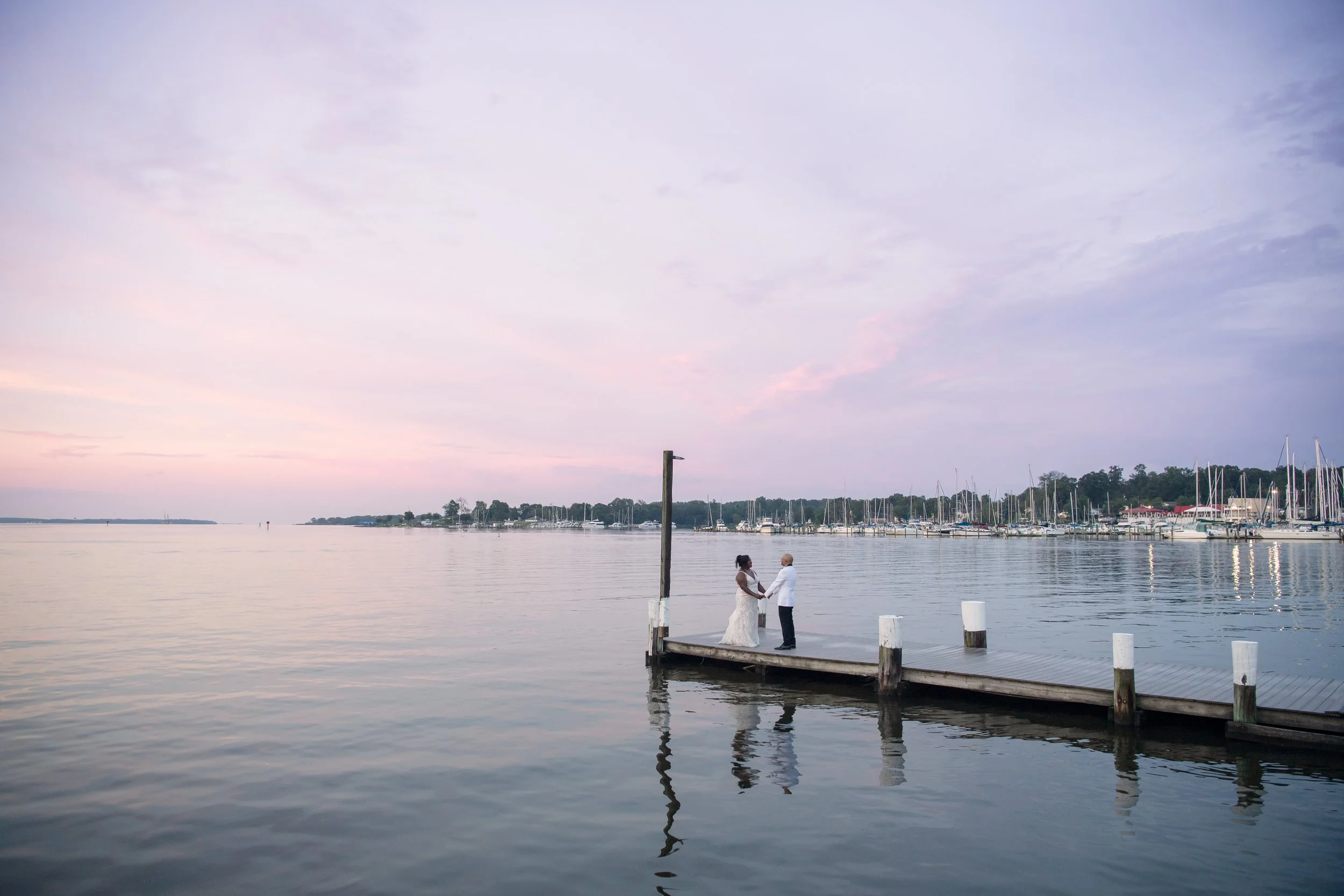 A couple standing on a dock, holding hands, at sunset near a marina with sailboats. The water is calm, and the sky has pastel pink and purple hues.