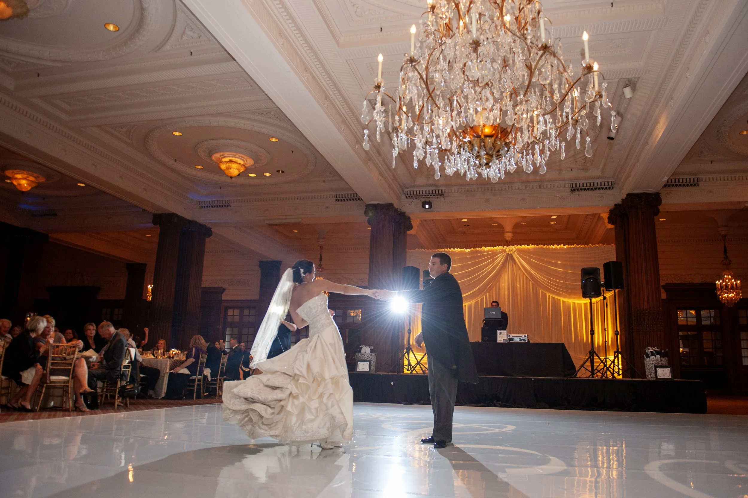 A bride and groom are dancing at their wedding reception in a grand ballroom with a chandelier overhead. Guests sit at tables watching the couple.