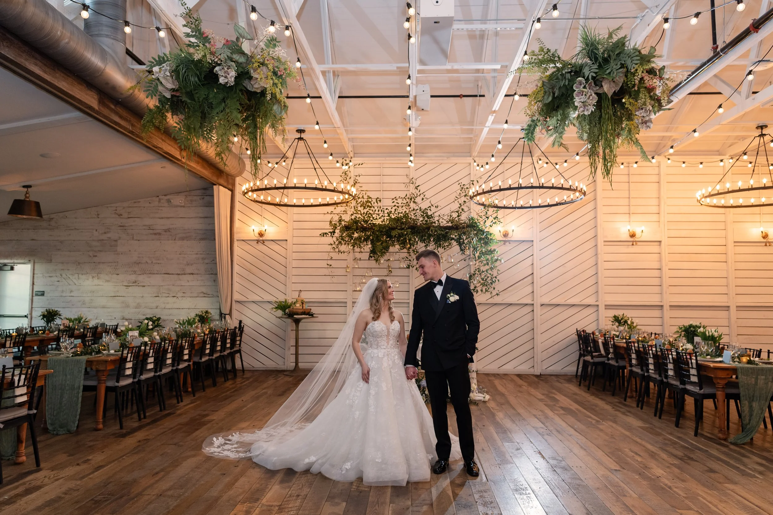 A bride and groom holding hands and smiling at each other during their wedding ceremony in a decorated venue with hanging lights and greenery.