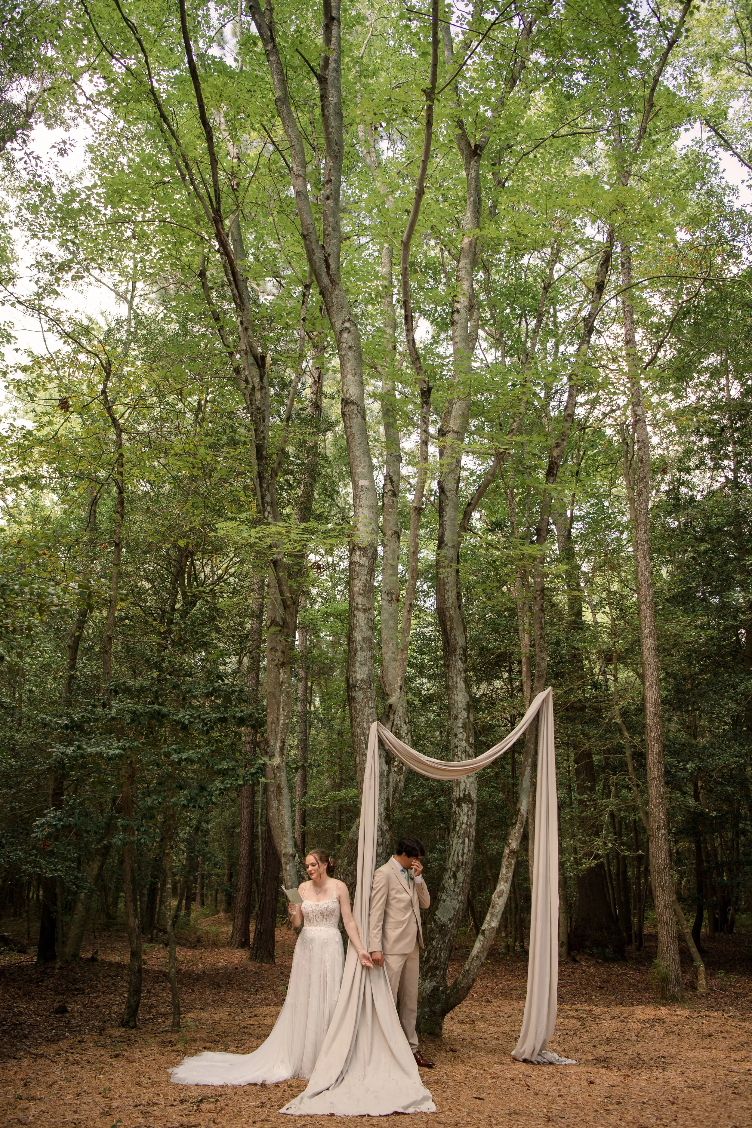 A bride and groom standing outdoors in a forest during their wedding ceremony. The bride is in a white wedding dress, holding a piece of paper, and smiling. The groom is in a beige suit, holding her hand, and appears emotional. A white fabric arch dr