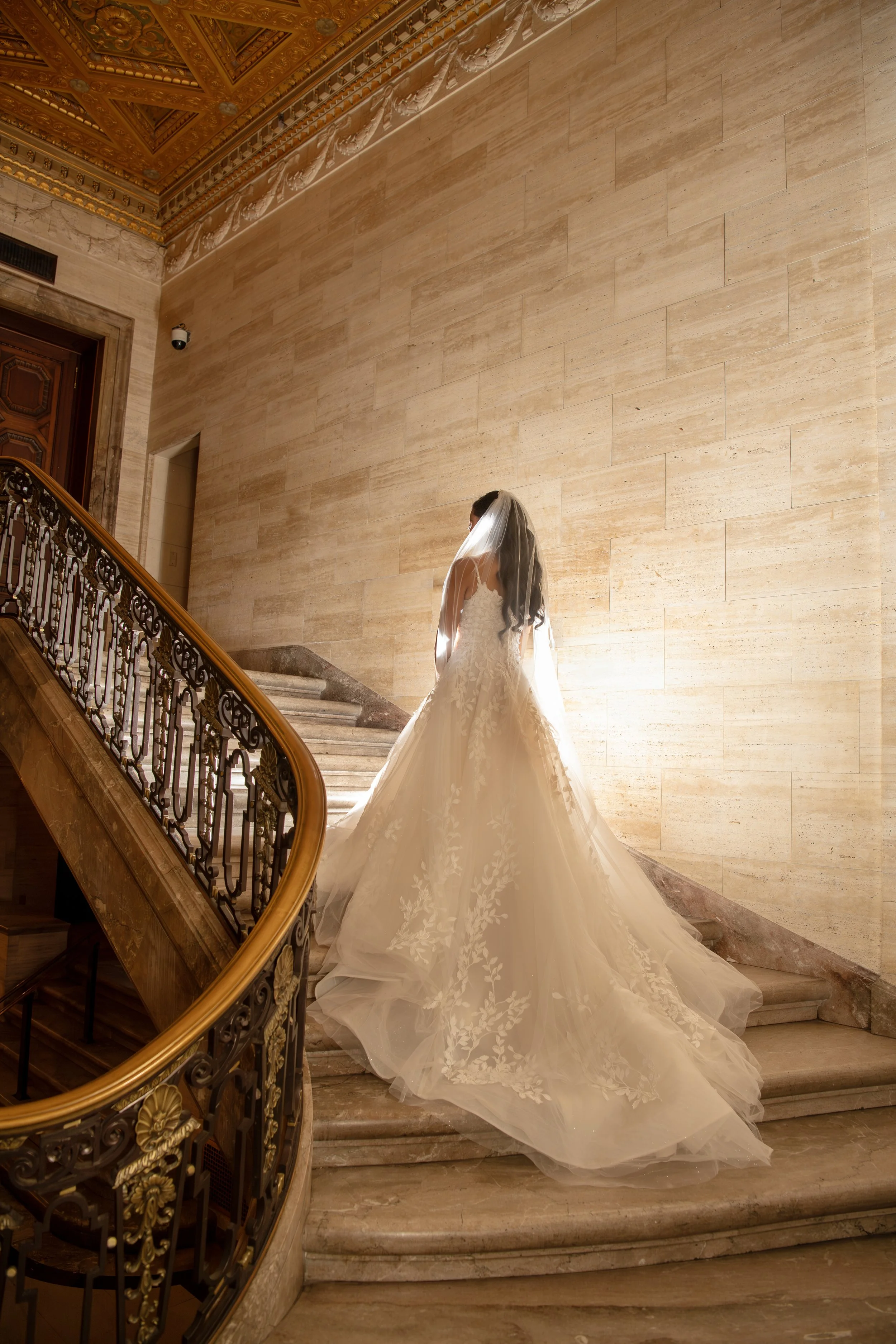 A bride in a white wedding gown with lace detail, standing on a grand staircase with ornate railing, in an elegant interior with marble walls and a decorated ceiling.