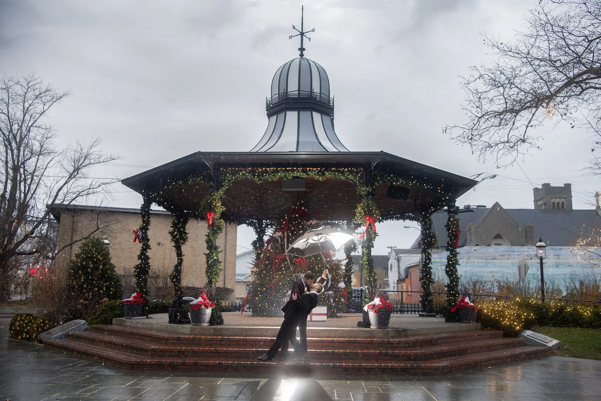 Couple dancing under an umbrella on a rainy day in a decorated festive park with Christmas lights and ornaments.
