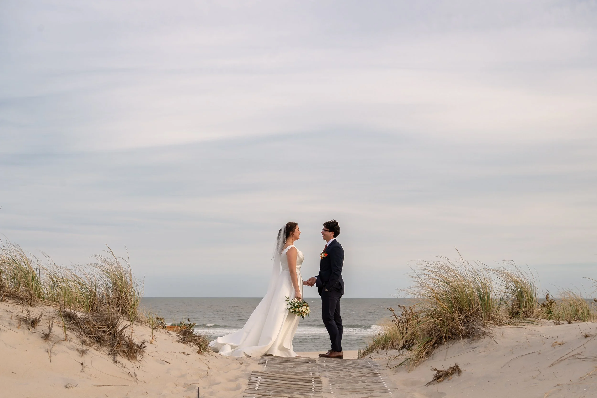 A bride and groom standing in the sand on a beach, holding hands and facing each other, with the ocean and cloudy sky in the background.