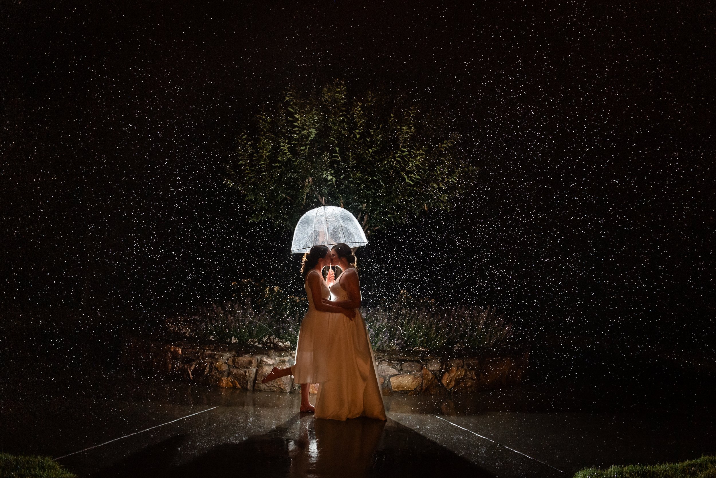 Two women in wedding dresses standing close under a transparent umbrella in the rain at night, with a tree and rain droplets illuminated in the background.
