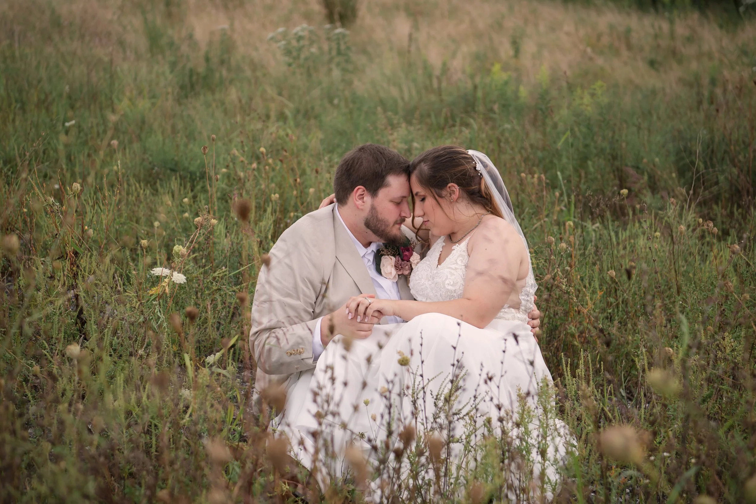 A newlywed couple sitting in a field of tall grass, touching foreheads and holding hands.