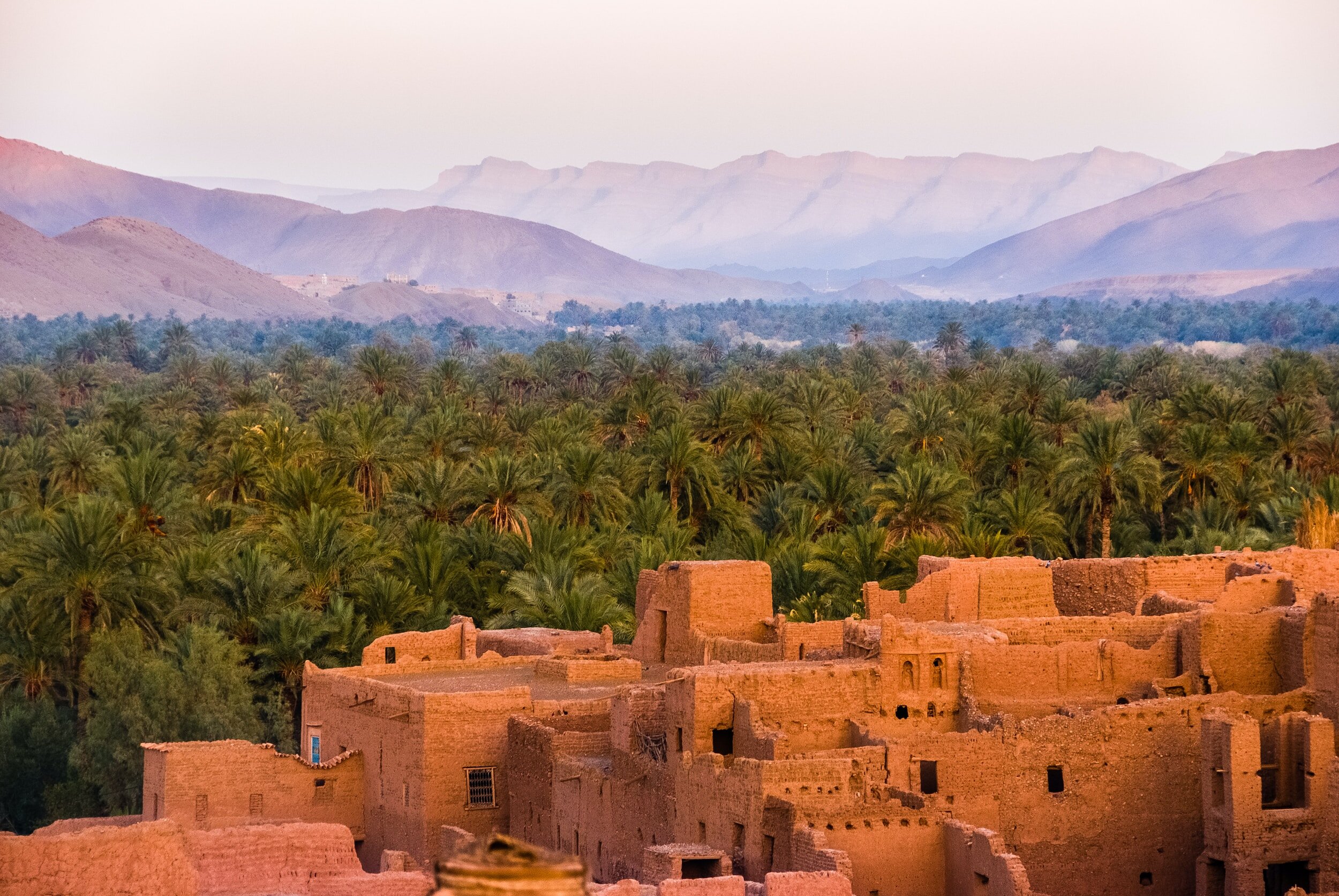 Ancient desert village with mud brick buildings in foreground, lush date palm oasis in middle, and distant mountains in background.